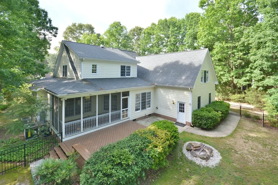 Two-story house with a screened porch, surrounded by trees and greenery, featuring a backyard with a fire pit and a deck.