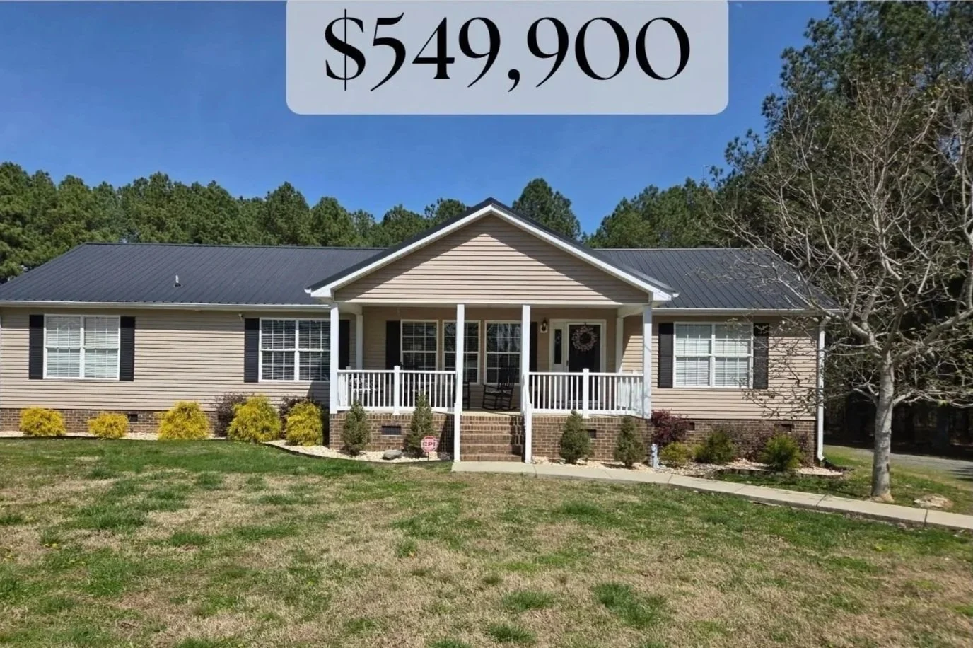 Front view of a beige single-story house with a metal roof, white porch, and garden with bushes and grass.