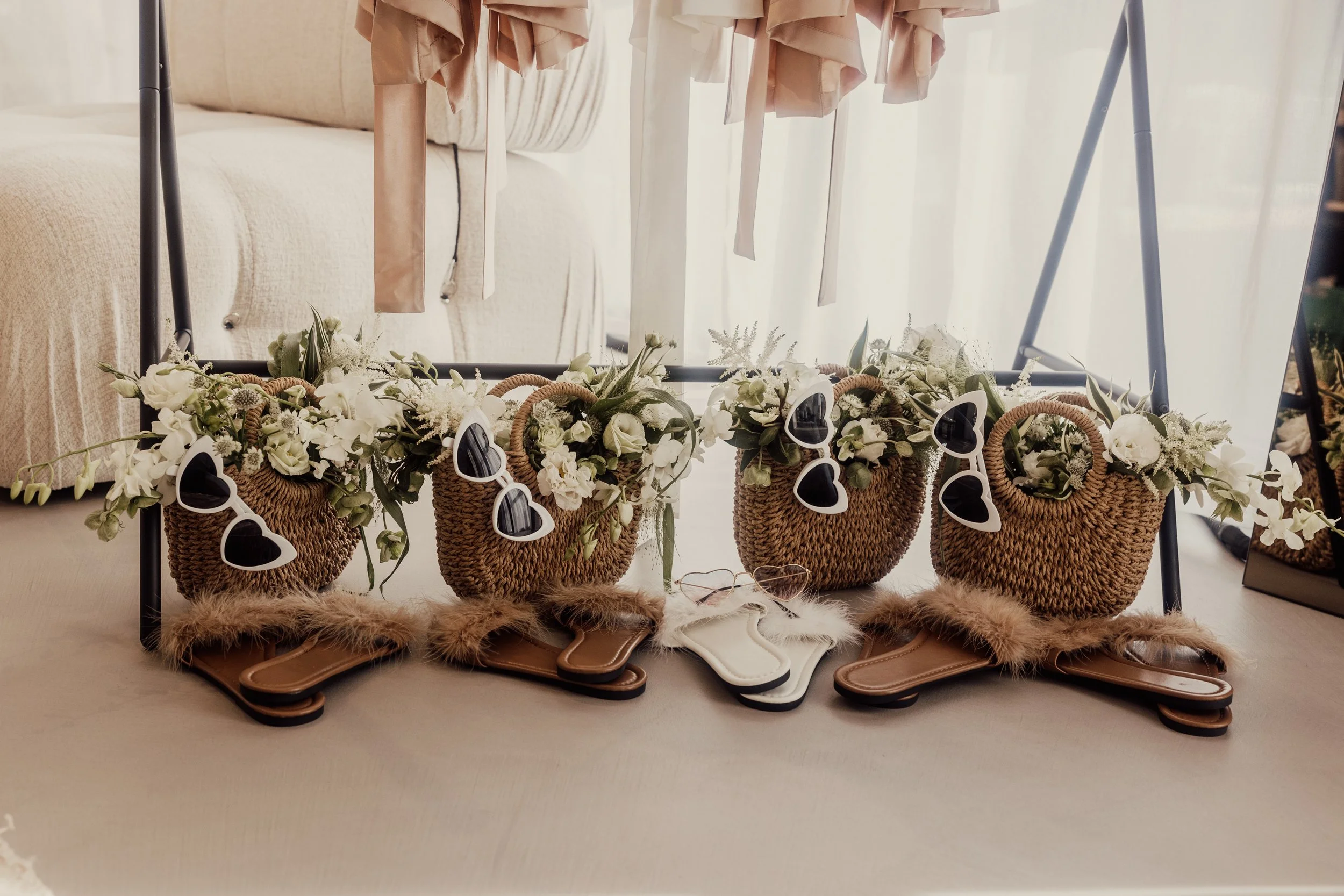 Four straw tote bags with flowers and heart-shaped sunglasses, paired with fluffy sandals, placed under hanging clothes indoors.