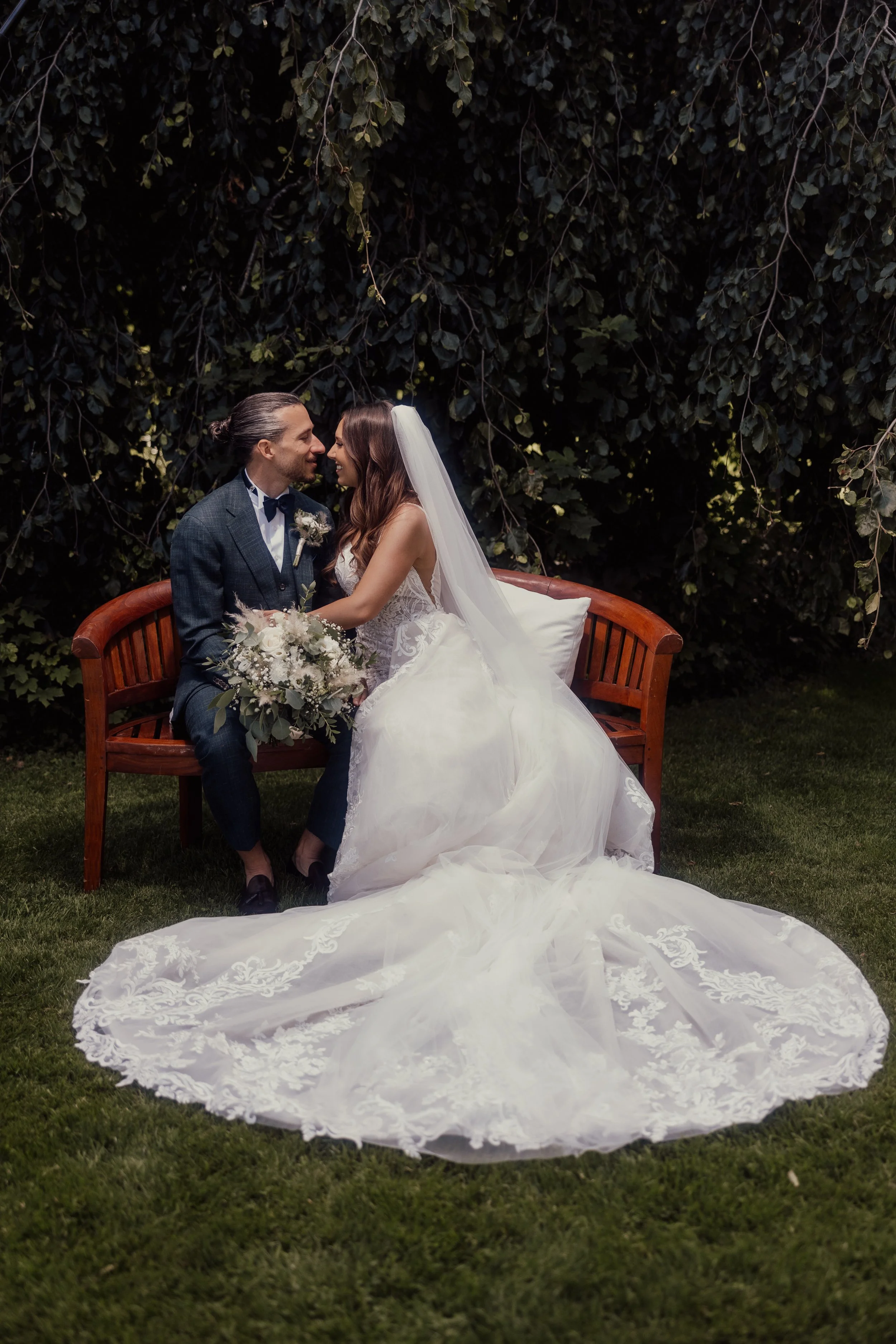 A bride and groom sitting on a wooden bench. The bride is wearing a white wedding dress with a long train and holding a bouquet. The groom is in a dark suit. They are sitting close together, surrounded by greenery.