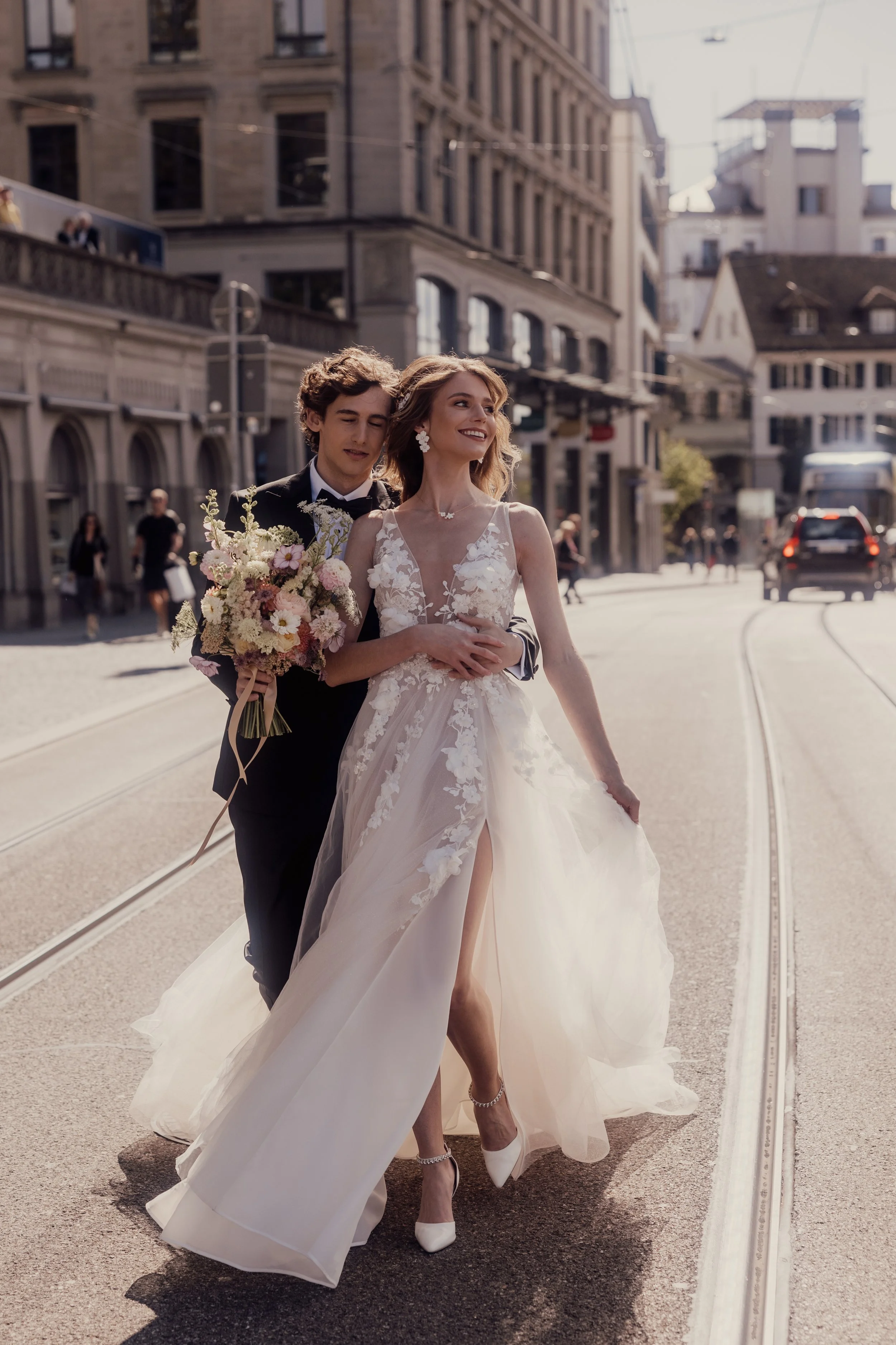 Bride and groom walking on a city street, bride holding a bouquet, wearing a white dress with floral details.