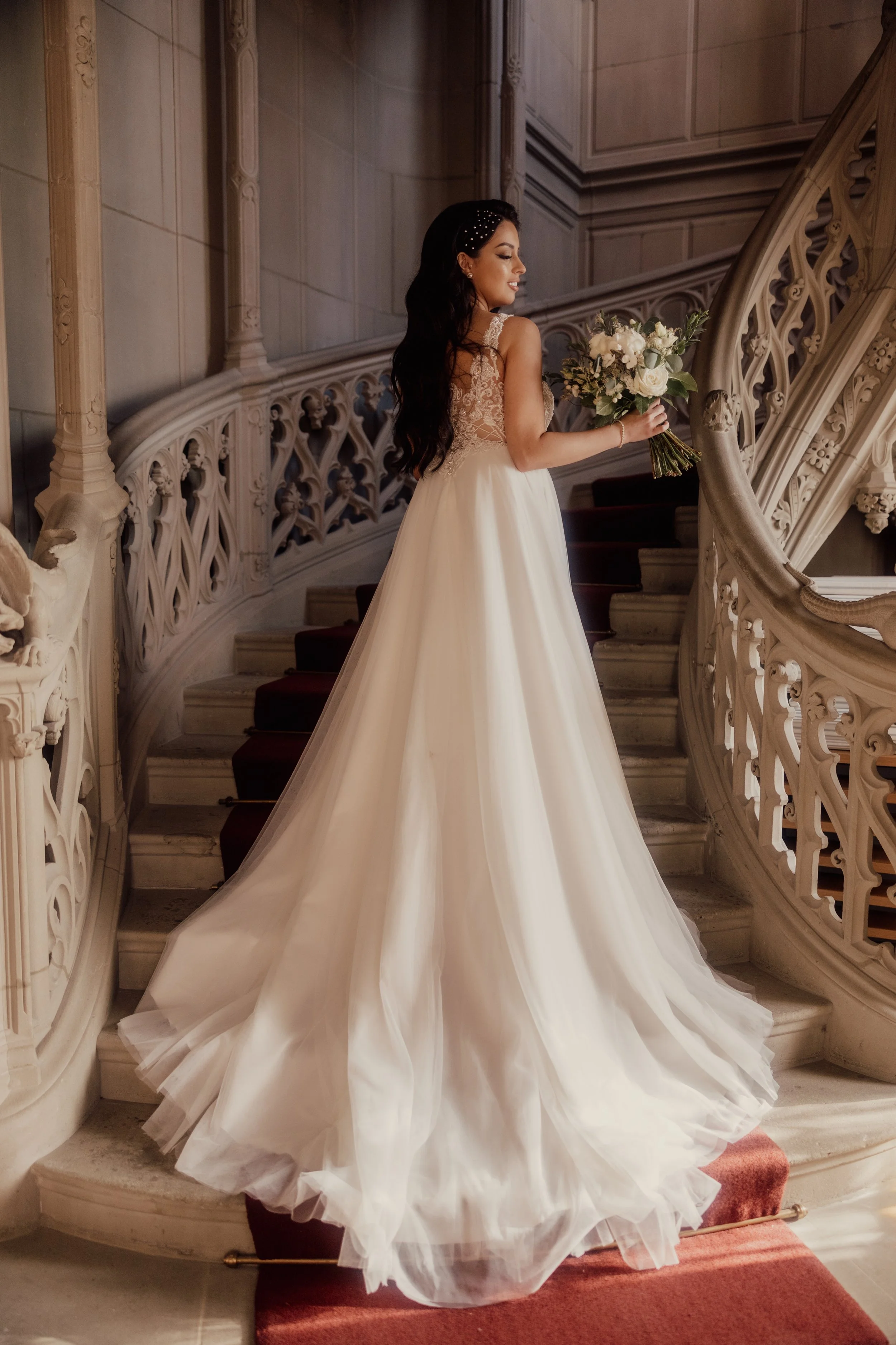 Bride in lace wedding dress holding bouquet on ornate staircase.