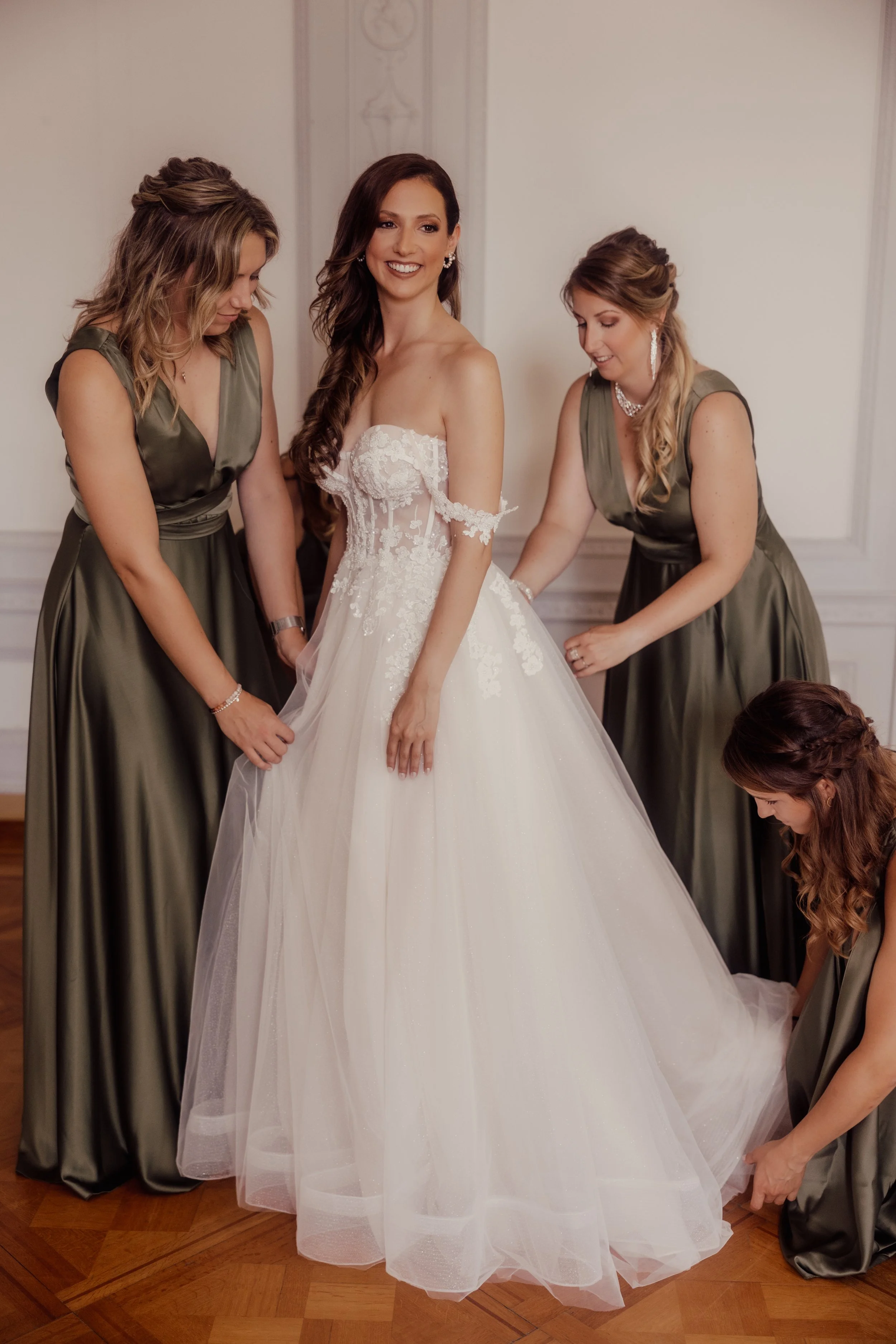 Bride in white wedding dress with three bridesmaids in olive green dresses helping her with gown adjustments.