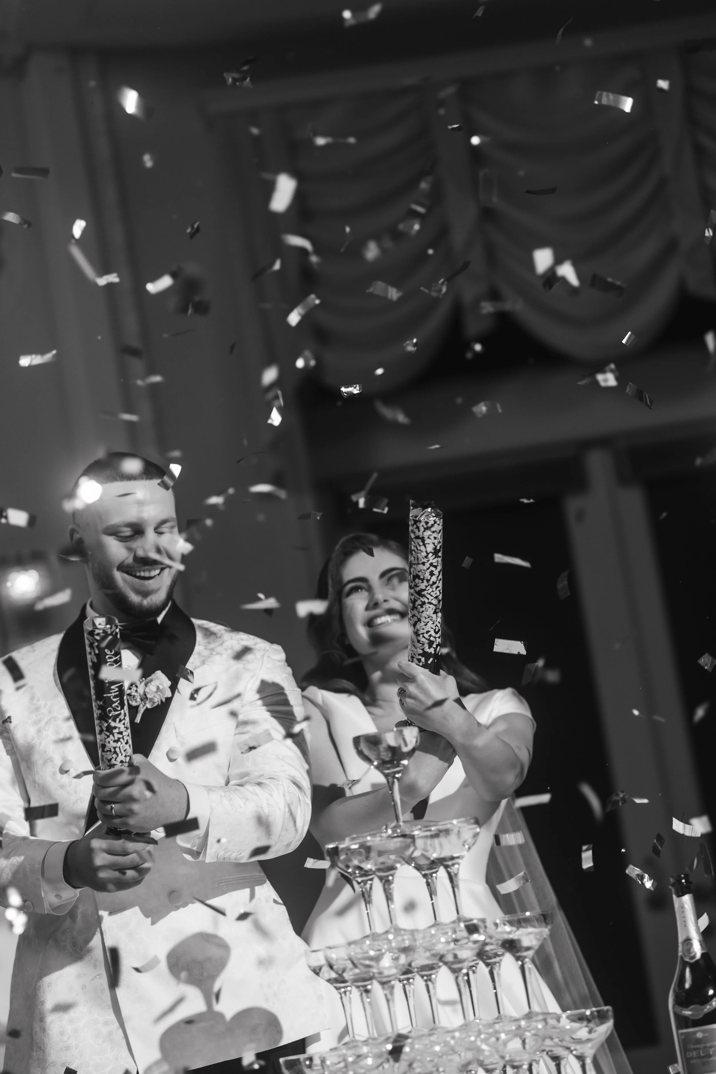 Black and white photo of a smiling couple celebrating, holding confetti tubes, near a champagne tower with falling confetti.