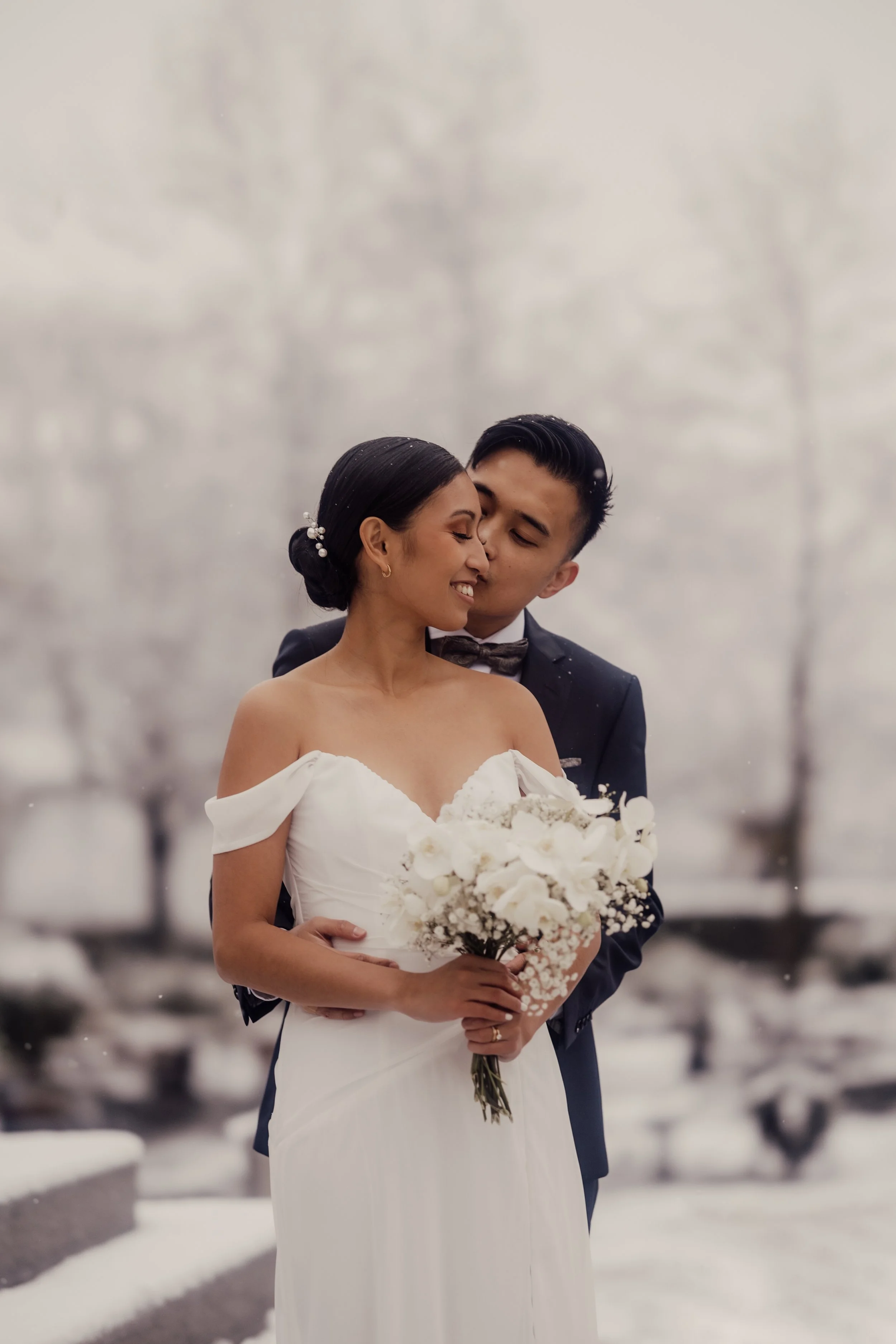 Bride and groom in snow, embracing, holding white bouquet, outdoor wedding scene.
