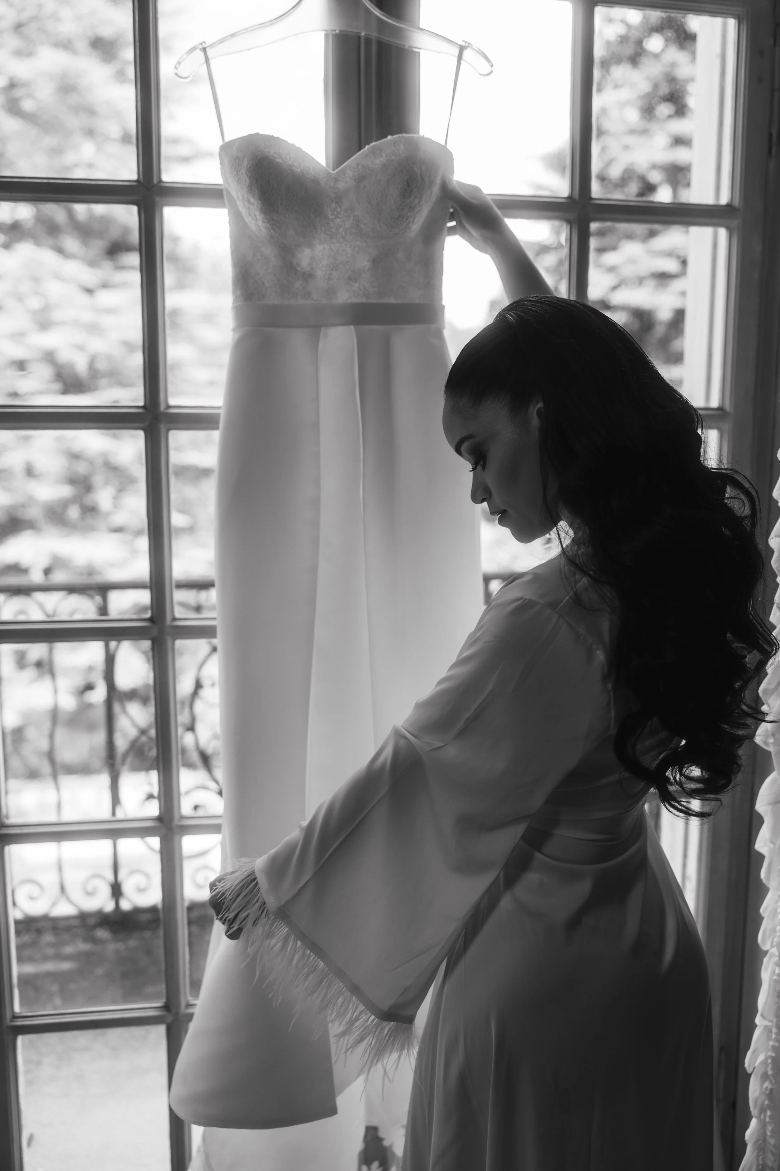 Black and white photo of a woman standing by a window, gazing at a wedding dress hanging on a hanger.