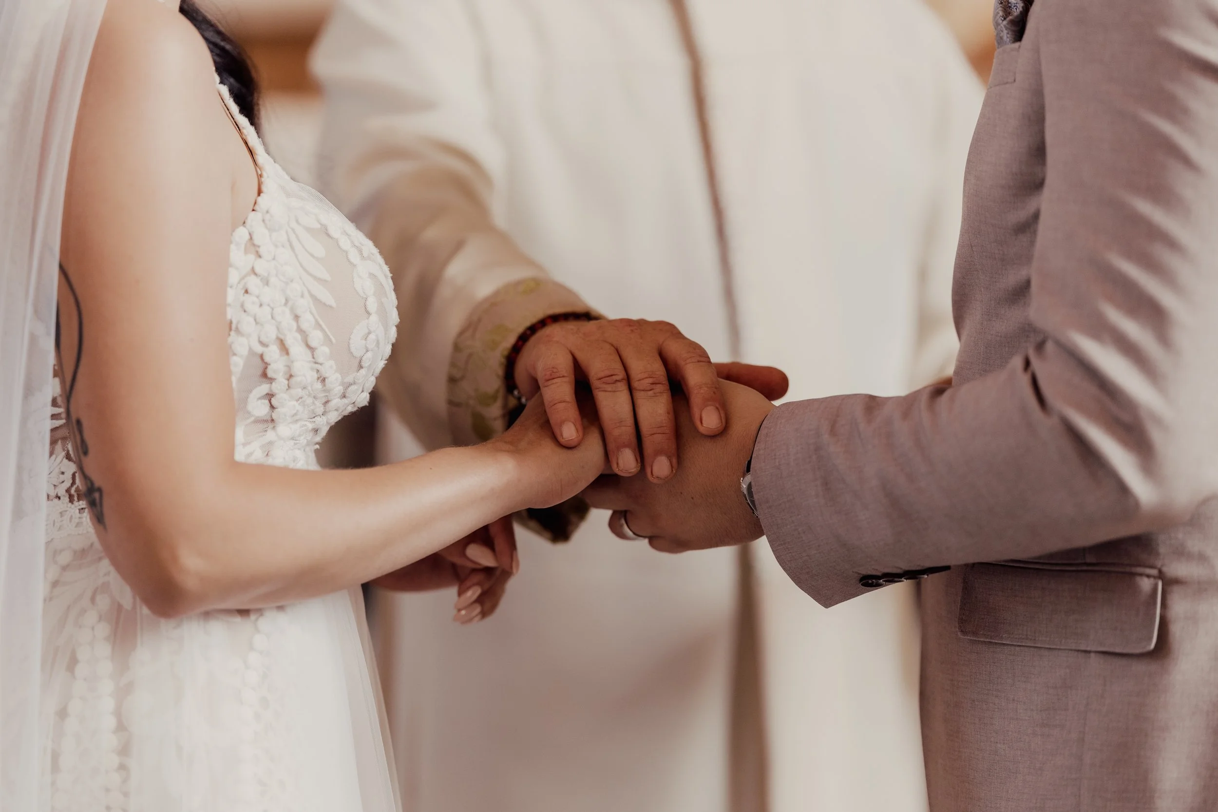 Bride and groom holding hands during wedding ceremony, with an officiant's hand placed over theirs.
