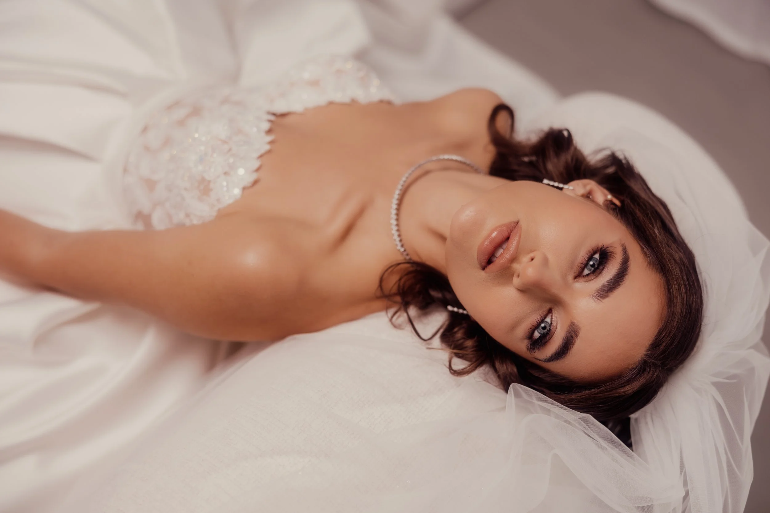 Bride lying down, wearing a strapless white wedding dress and a pearl necklace, looking at the camera.
