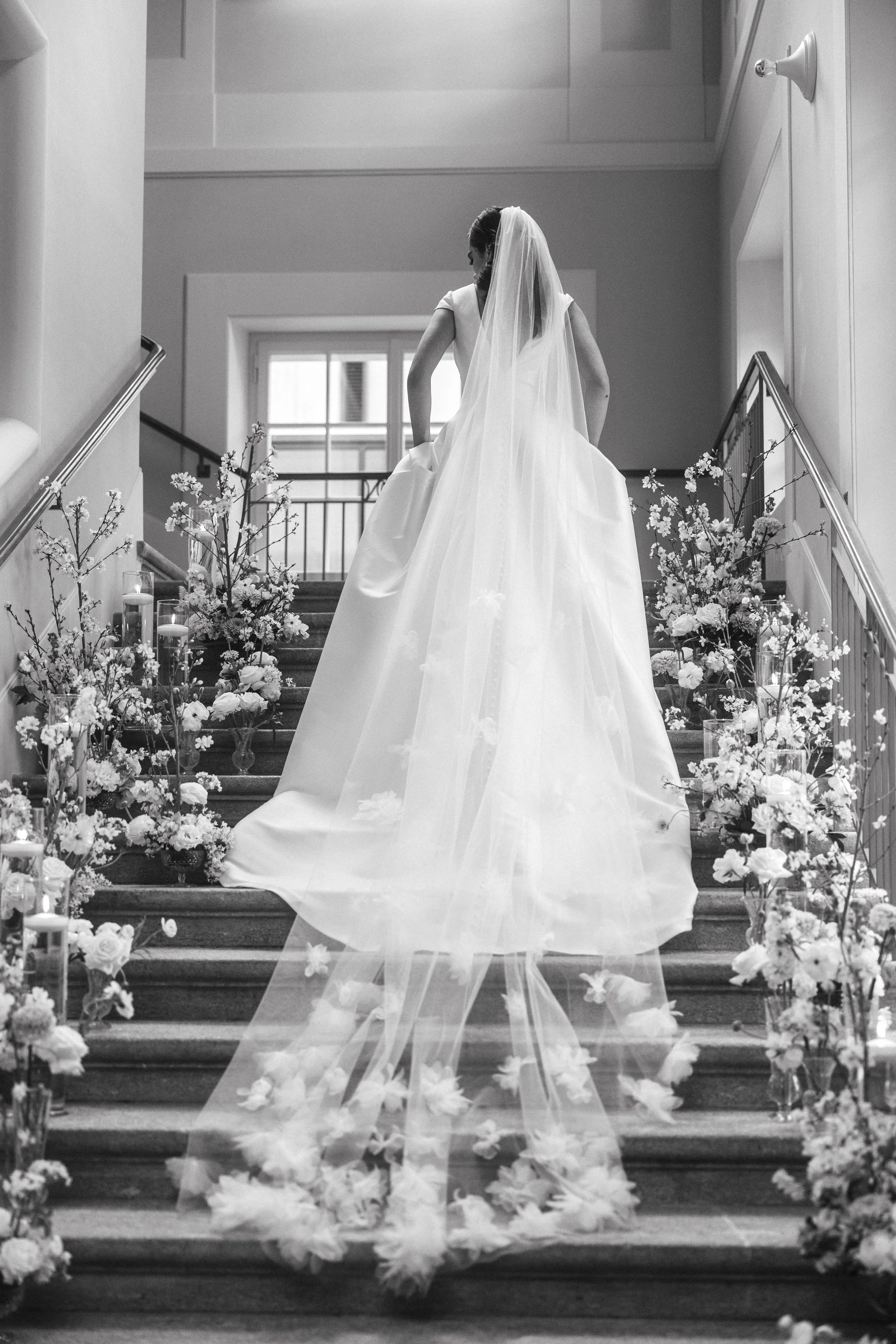 Bride walking up staircase adorned with flowers, wearing a long veil and gown.