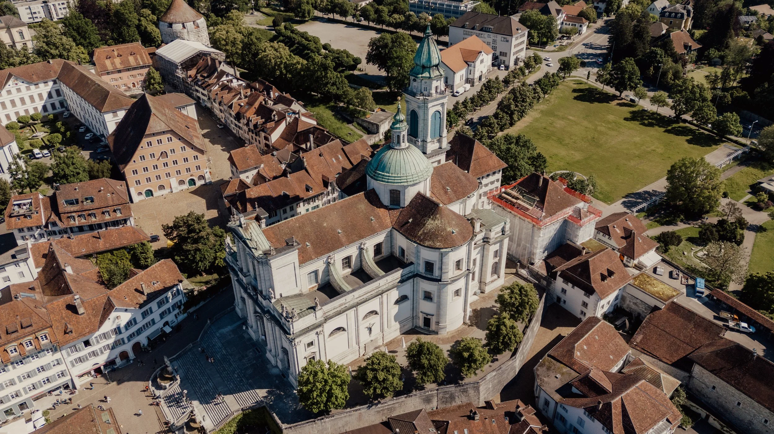 Aerial view of a historic city with a prominent cathedral featuring a green dome and clock tower, surrounded by traditional European architecture with red tile roofs, trees, and open spaces.