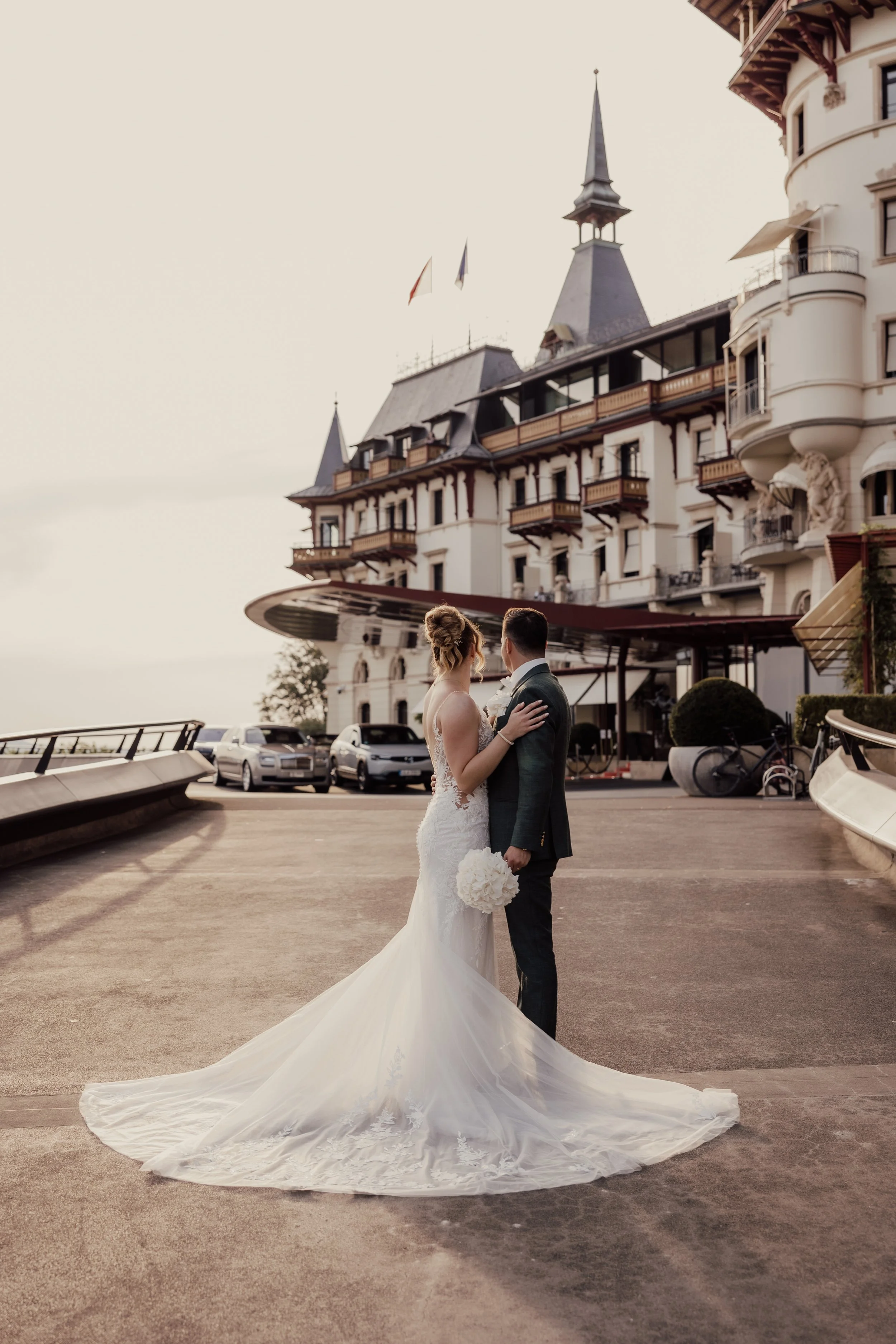 Bride and groom embracing in front of a grand hotel with ornate architecture and flags.
