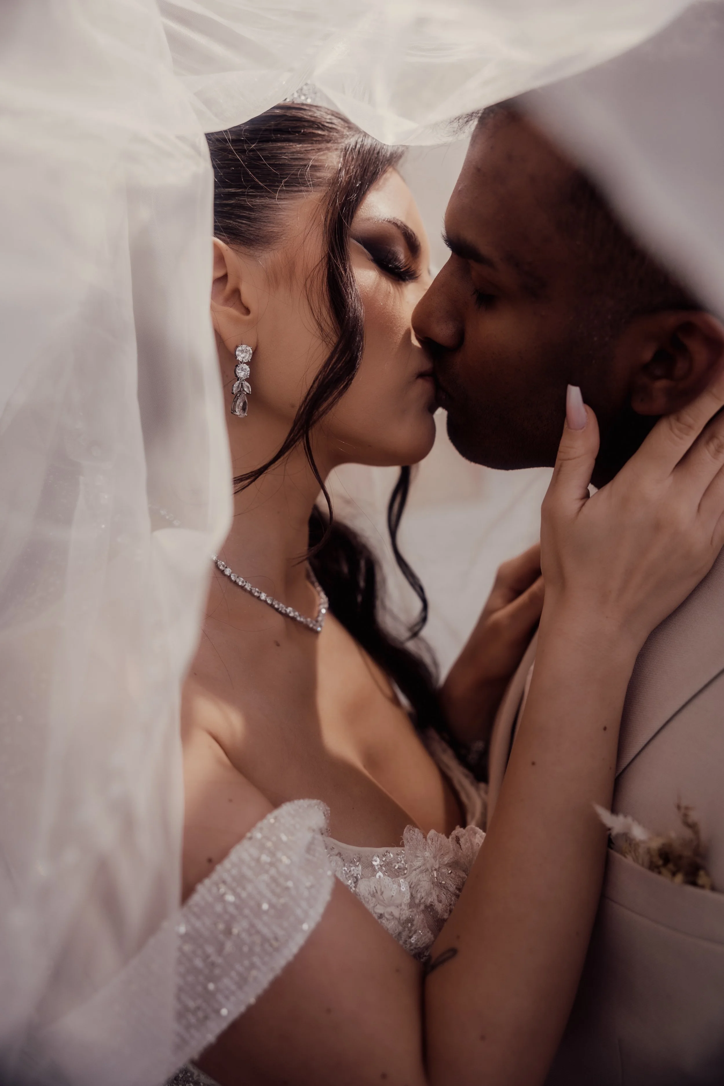 A bride and groom kissing under a veil, with the bride wearing diamond earrings and necklace.