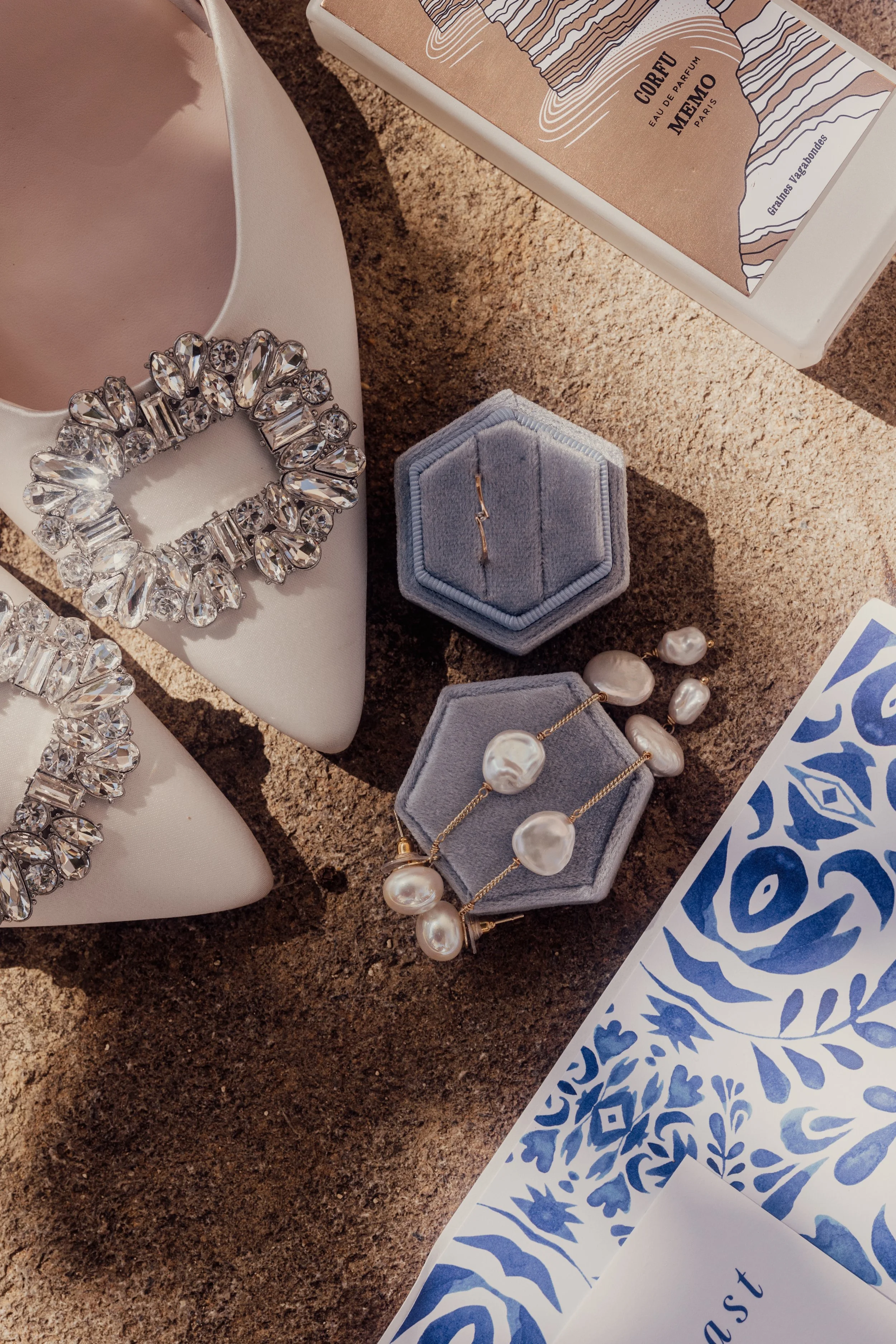Bridal flat shoes with jeweled buckles, pearl jewelry, a blue velvet ring box, a perfume box, and blue floral pattern paper on a textured surface.