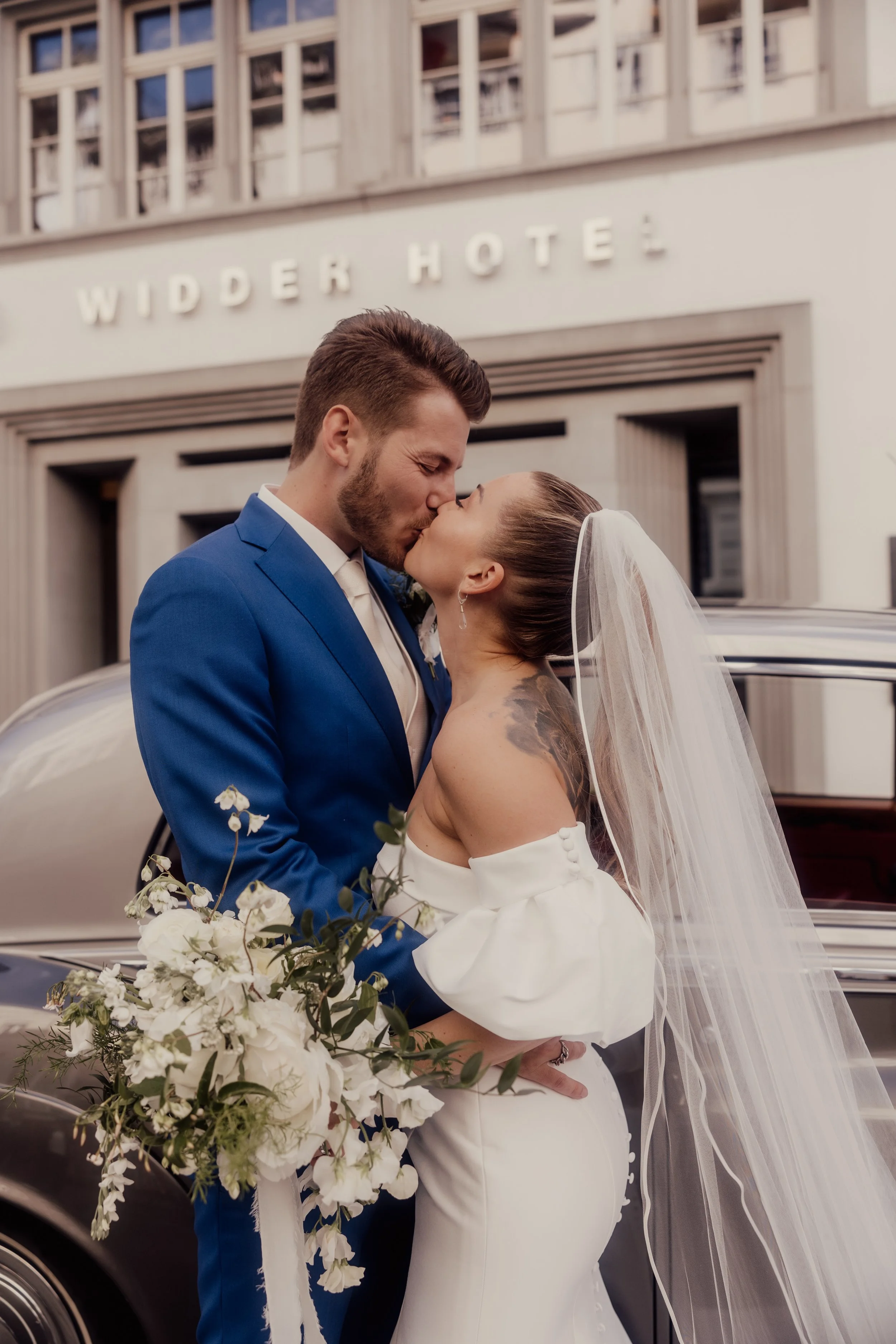 Bride and groom kissing in front of a hotel, bride in white dress holding bouquet, groom in blue suit.