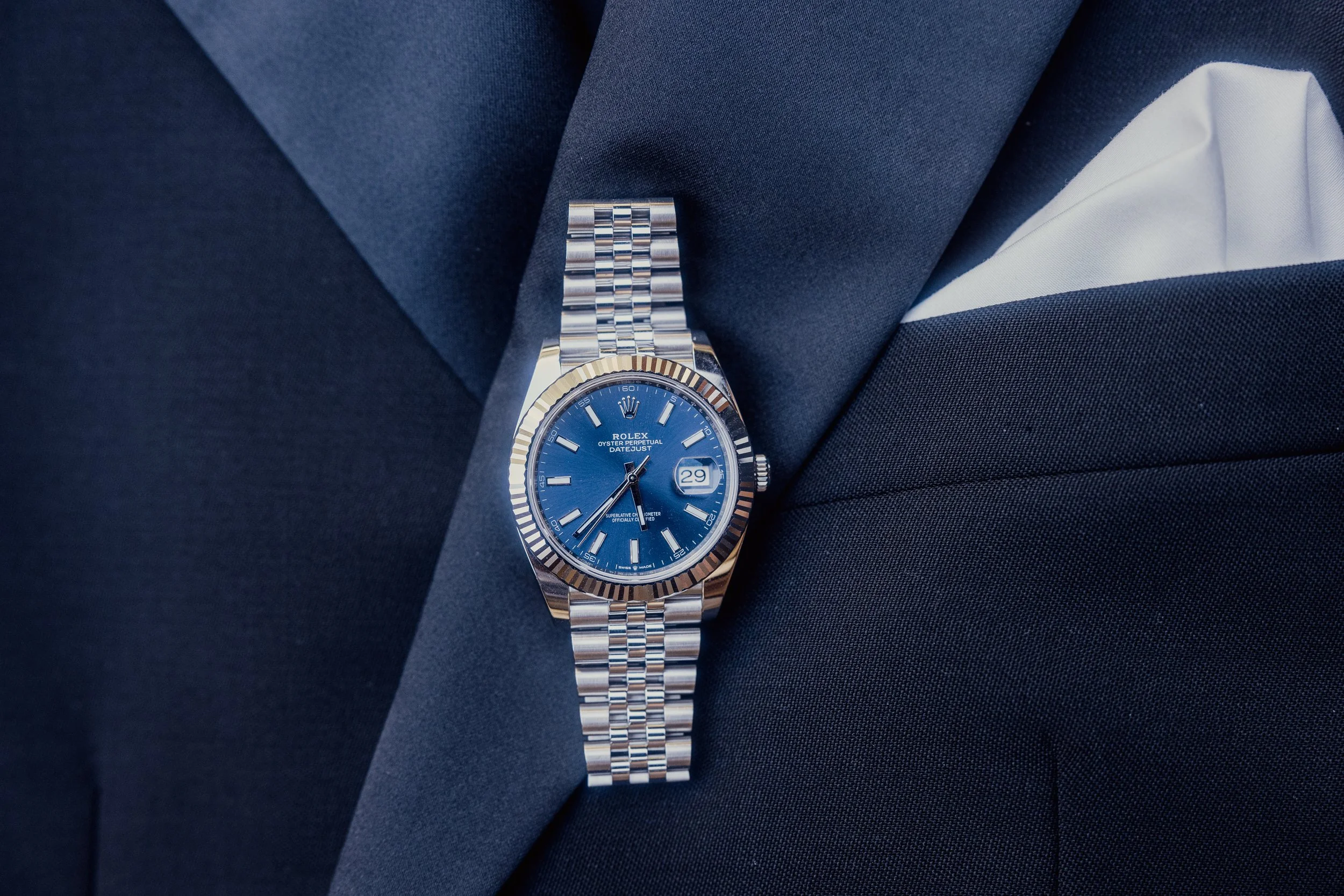 Close-up of a luxury watch with a blue dial and silver bracelet, resting on a dark suit with a white pocket square.