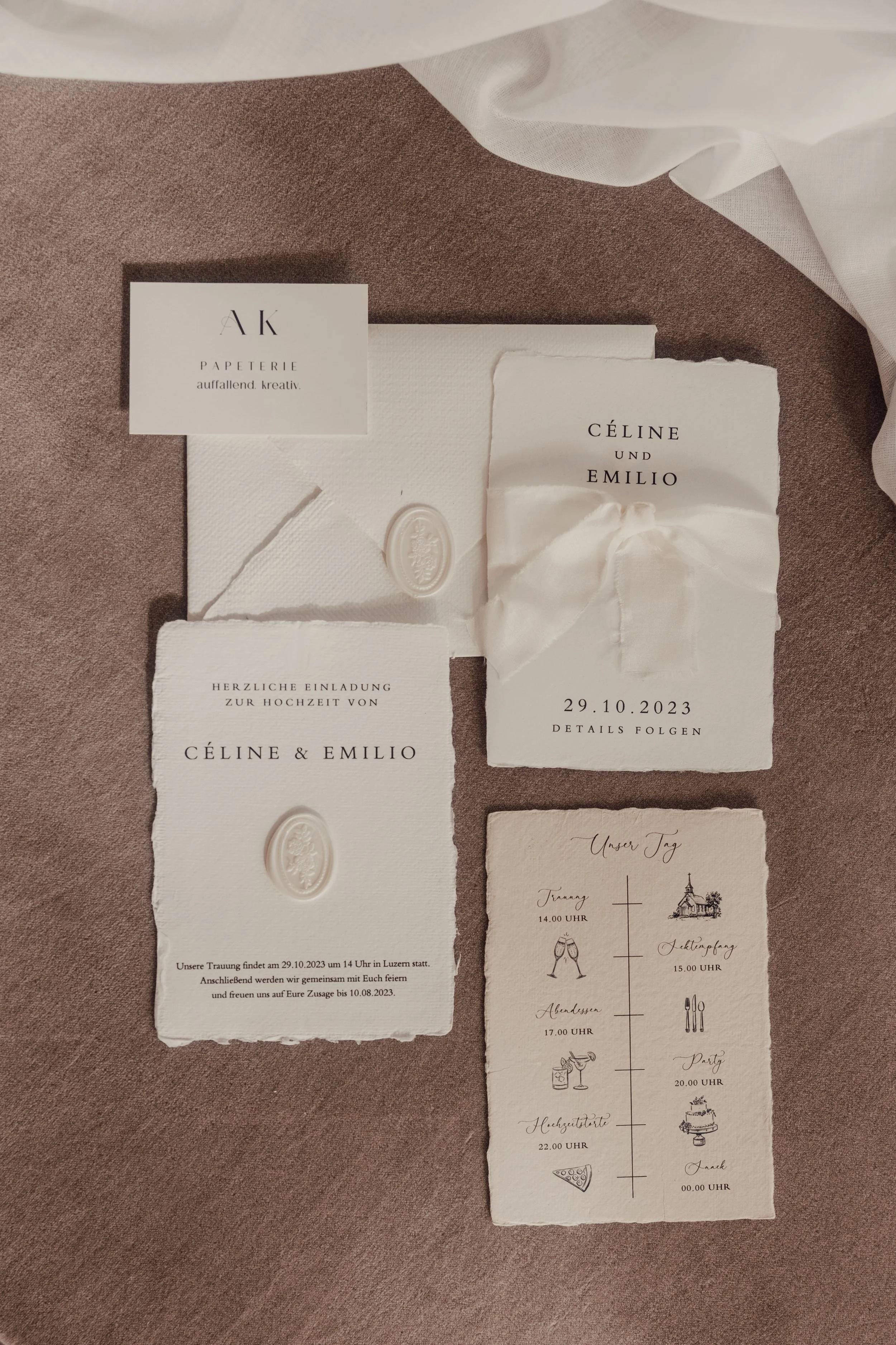 Elegant wedding invitation set for Céline and Emilio with cream-colored cards featuring embossed details and a timeline of wedding events. Includes a business card for AK Papeterie and soft fabric ribbon accents.