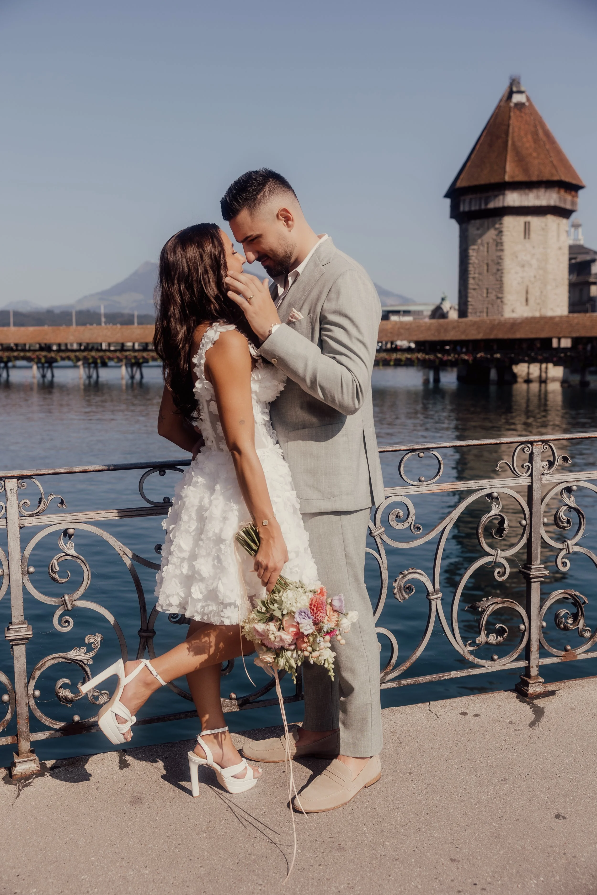 Couple embracing on a bridge with a tower in the background, both dressed in wedding attire, woman holding a bouquet.