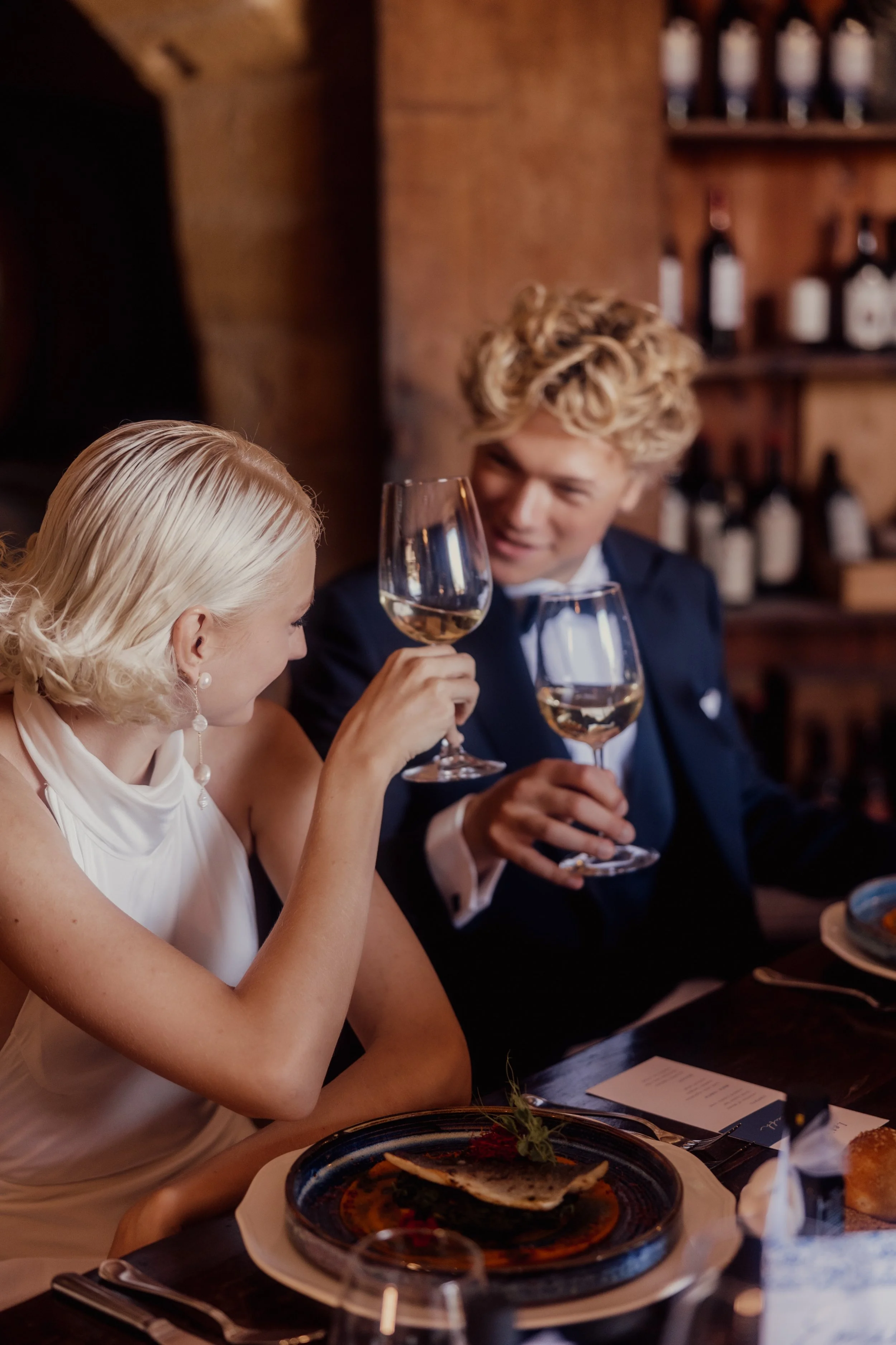 A couple dressed elegantly, with a man in a suit and a woman in a white dress, toasting with glasses of white wine at a restaurant table.