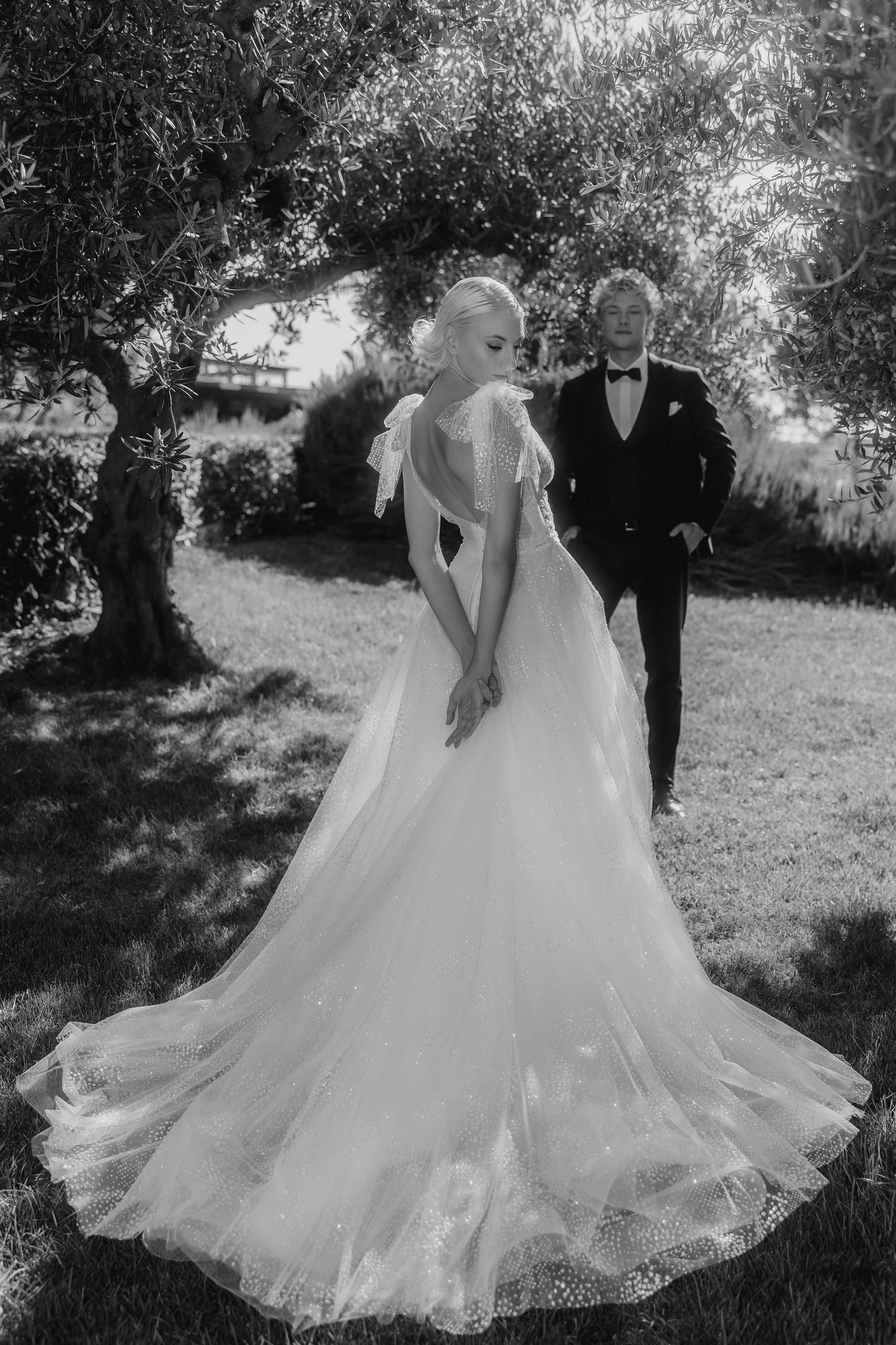 Bride in a wedding dress with a groom in a tuxedo standing outdoors under a tree.