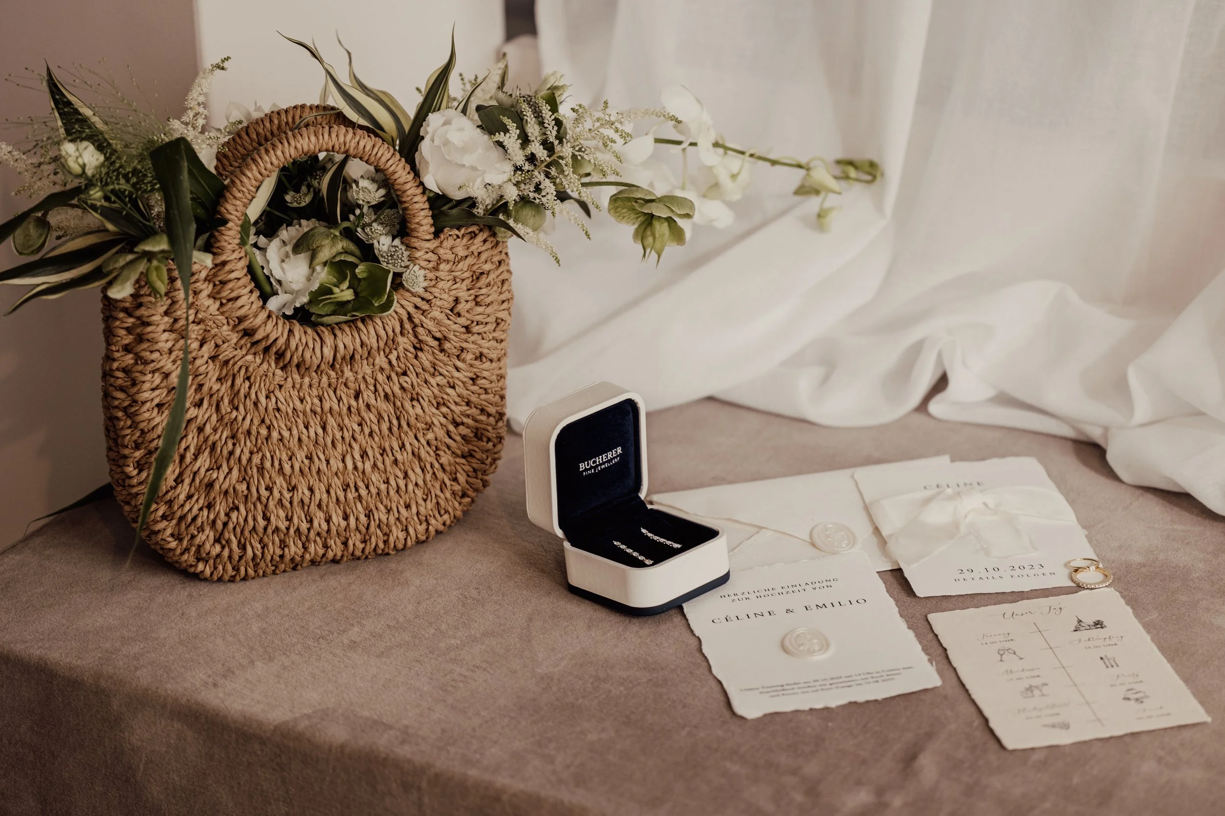 Wedding arrangement with a straw bag filled with flowers, a ring box, and wedding invitations on a table.