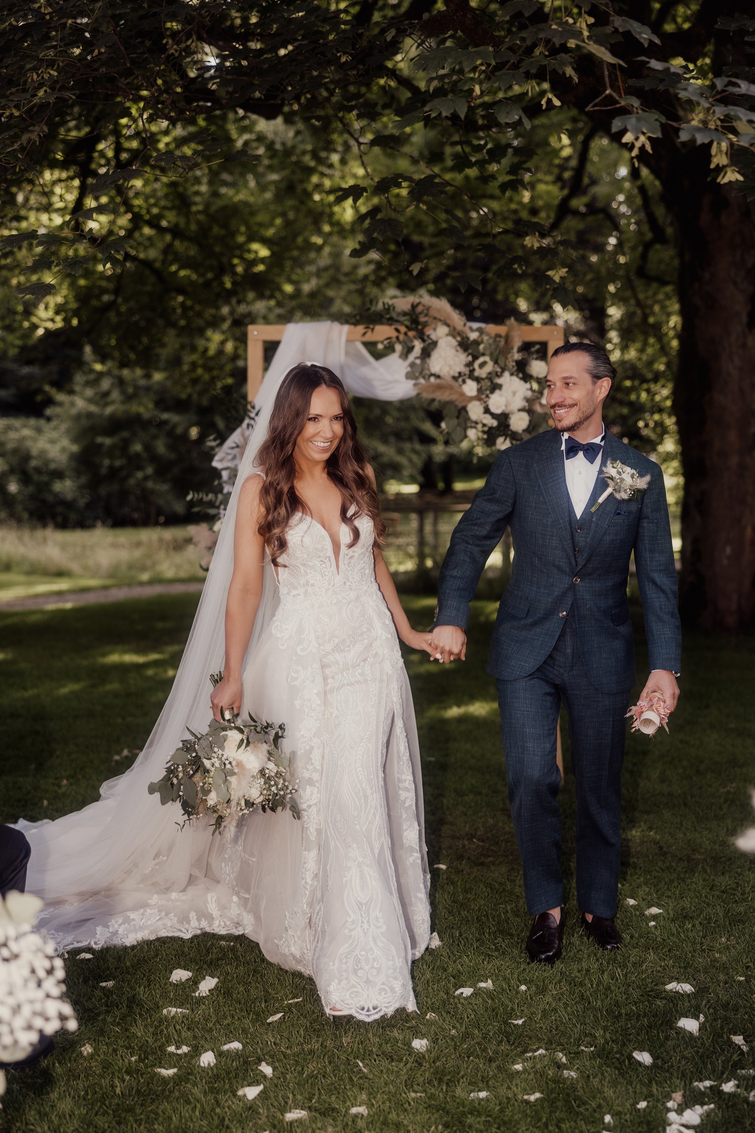 Bride and groom holding hands and smiling outdoors, dressed in wedding attire, with greenery in the background.