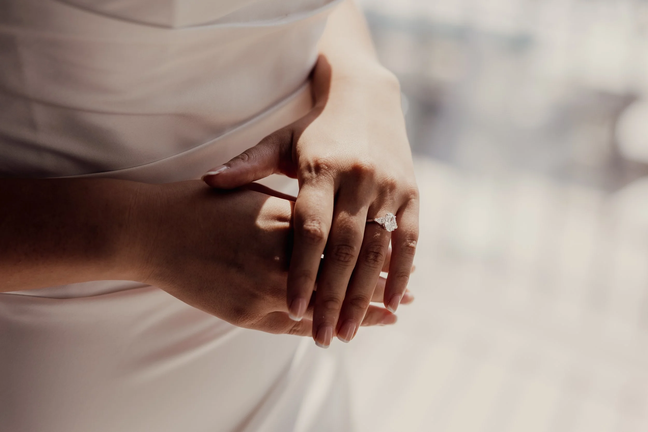 Close-up of hands with an engagement ring on a finger, wearing a white dress.