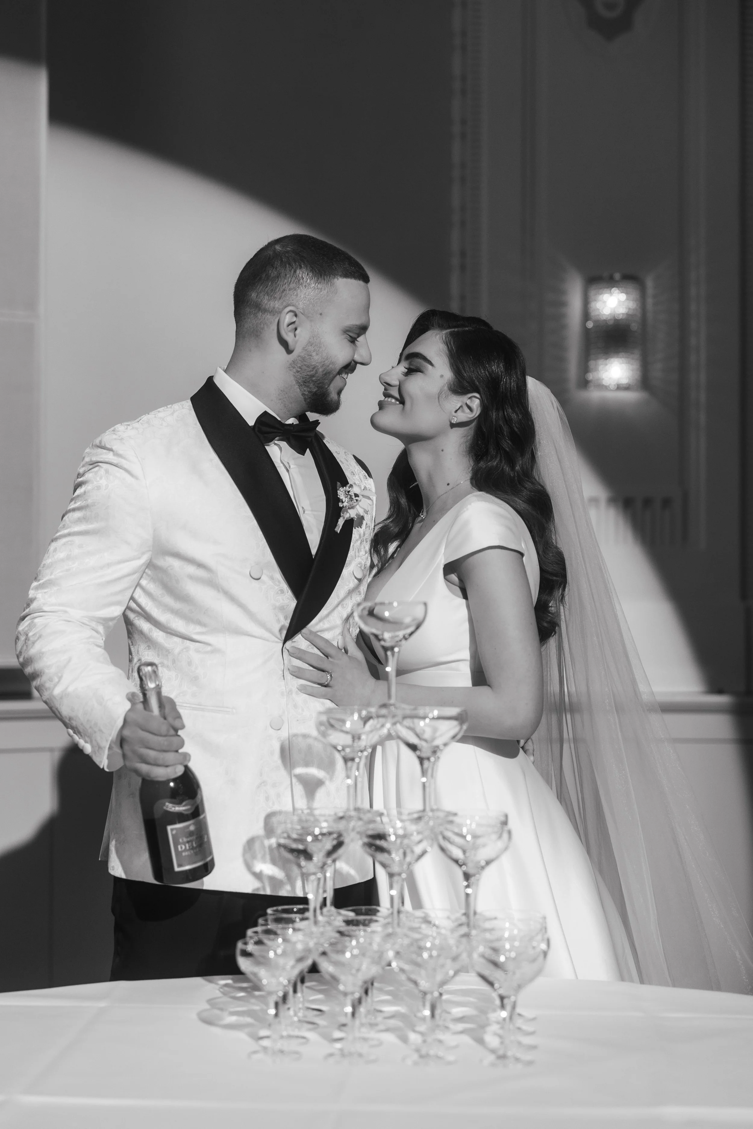 Bride and groom standing next to a champagne tower, groom holding a bottle, looking at each other, in black and white.