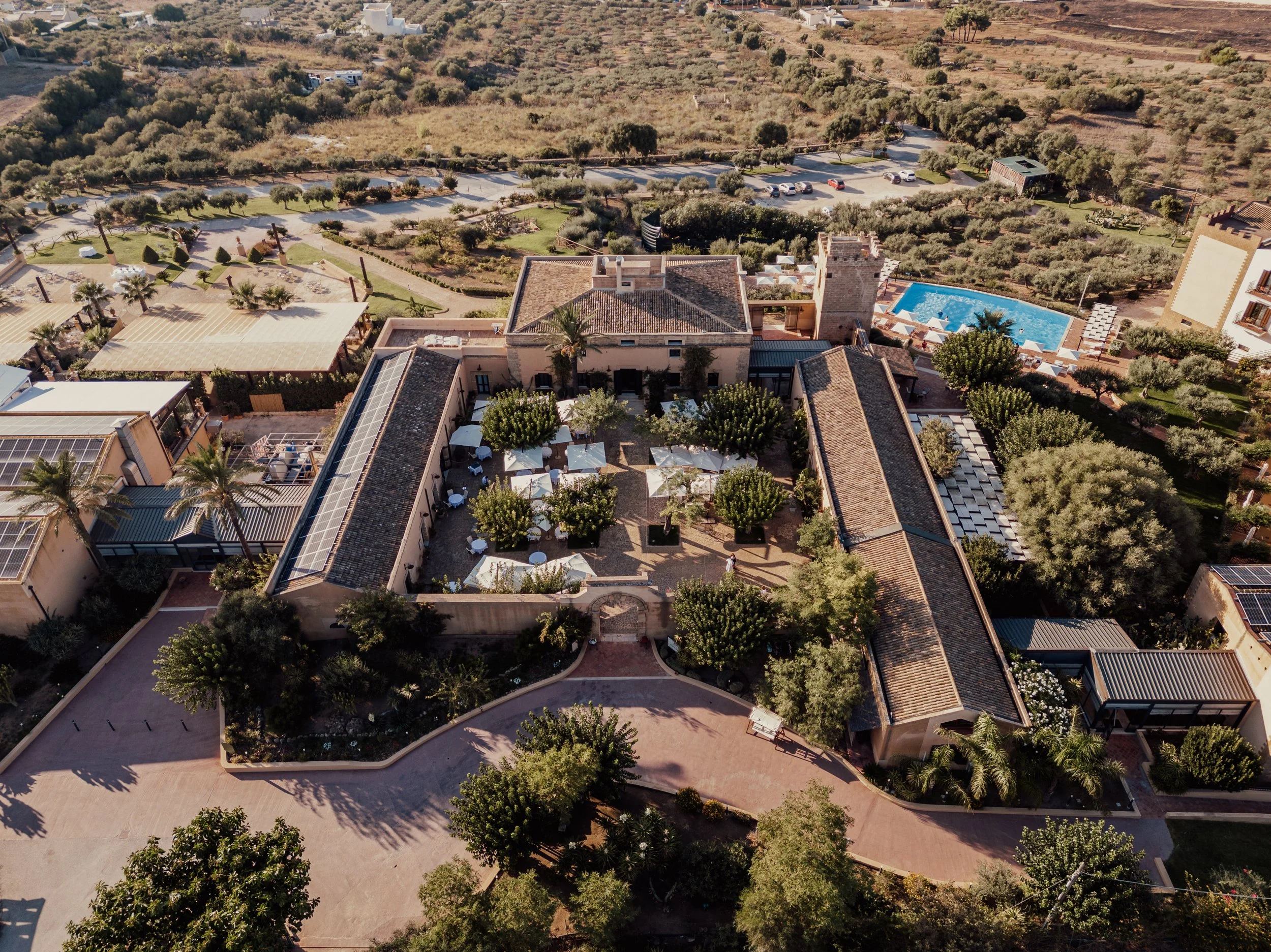 Aerial view of a Mediterranean-style villa surrounded by gardens, trees, and a swimming pool, with solar panels on the roof. The landscape includes parking areas and pathways, with fields and vegetation extending into the background.