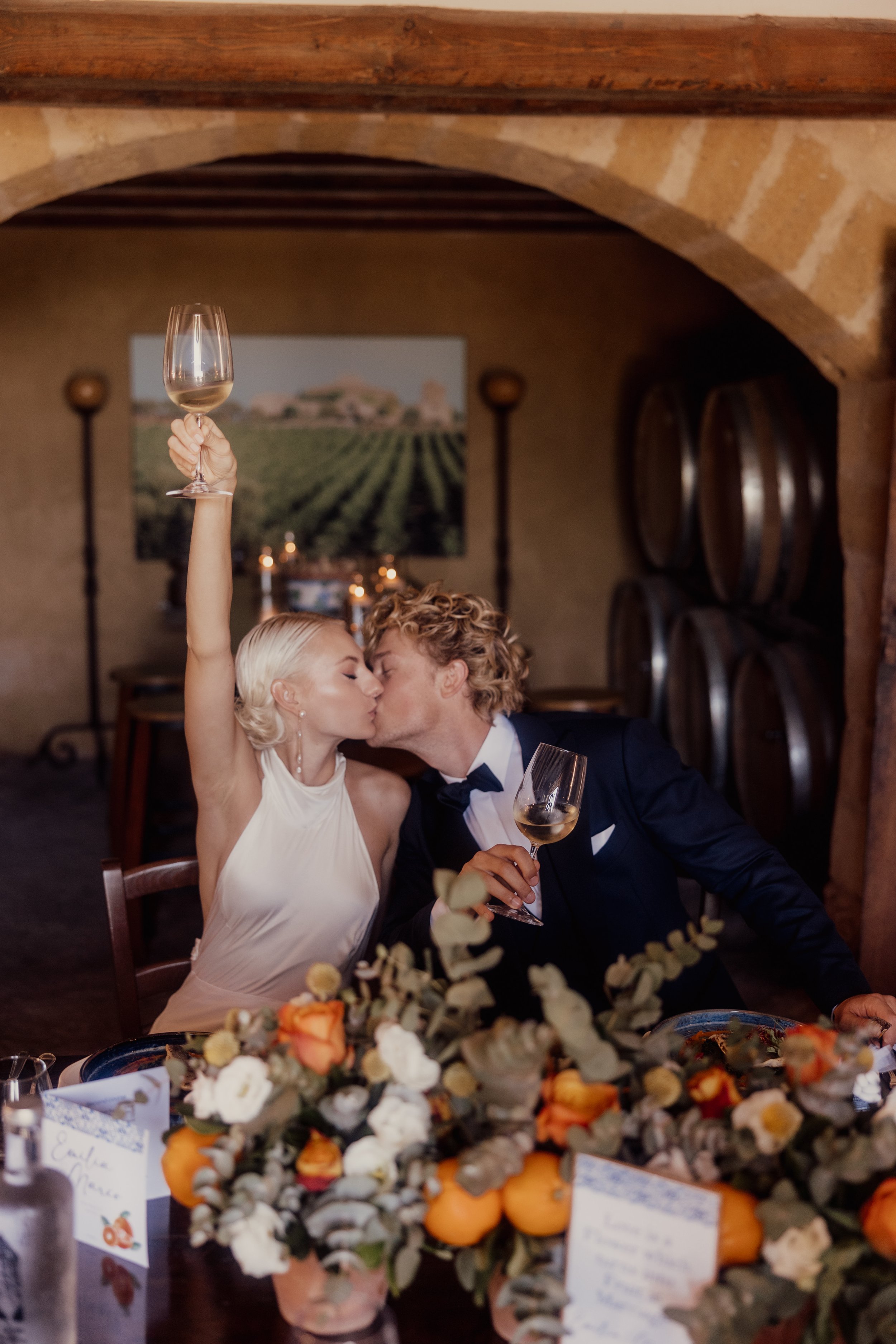 A couple kissing at a decorated dining table with wine glasses raised, surrounded by flowers and wine barrels in the background.