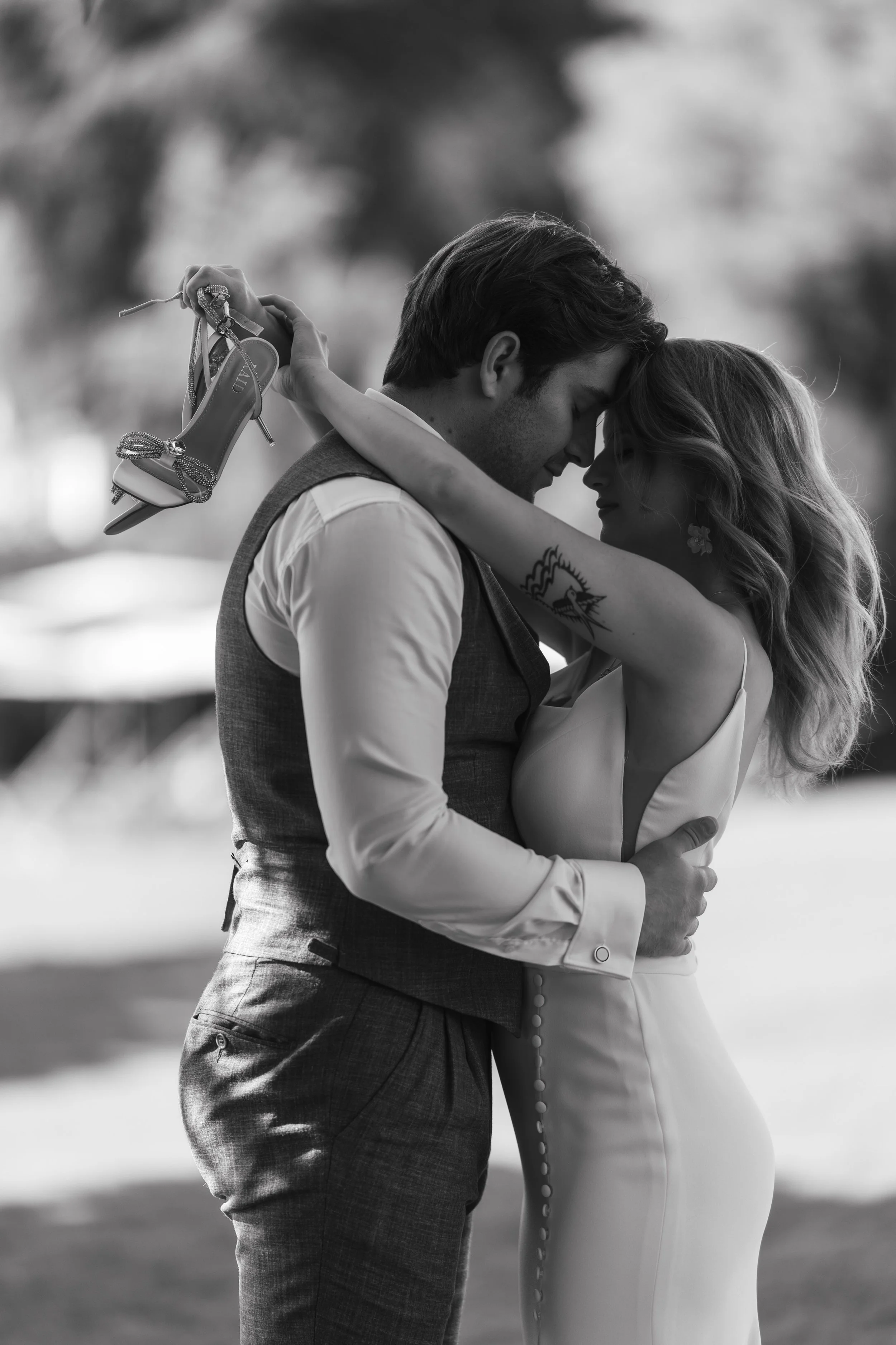A couple embracing in an intimate pose, with the woman holding high-heeled shoes over the man's shoulder. The setting appears to be outdoors, with a blurred background, in black and white.