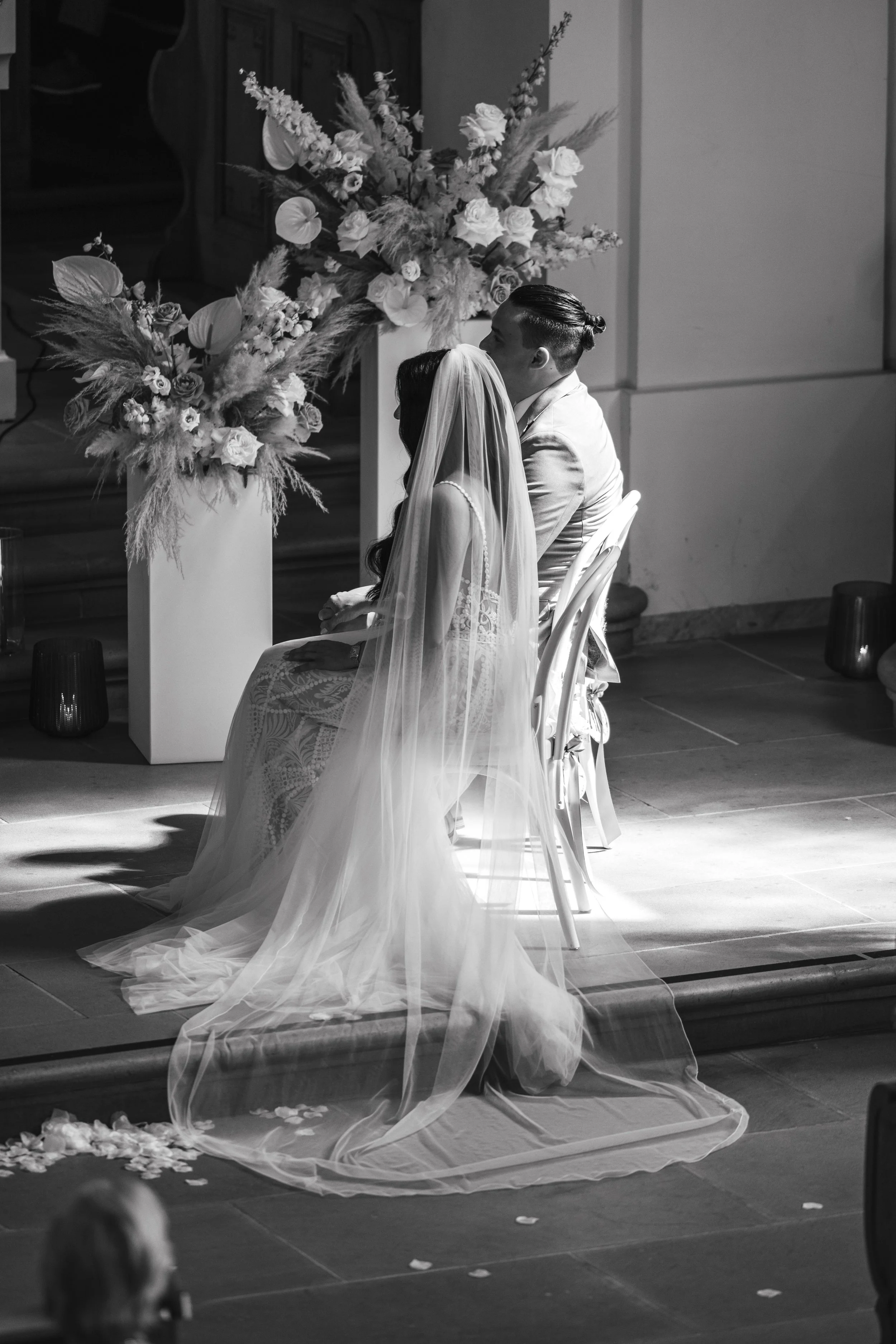 Bride and groom seated during a wedding ceremony, surrounded by floral arrangements, with the bride wearing a veil and the scene in black and white.