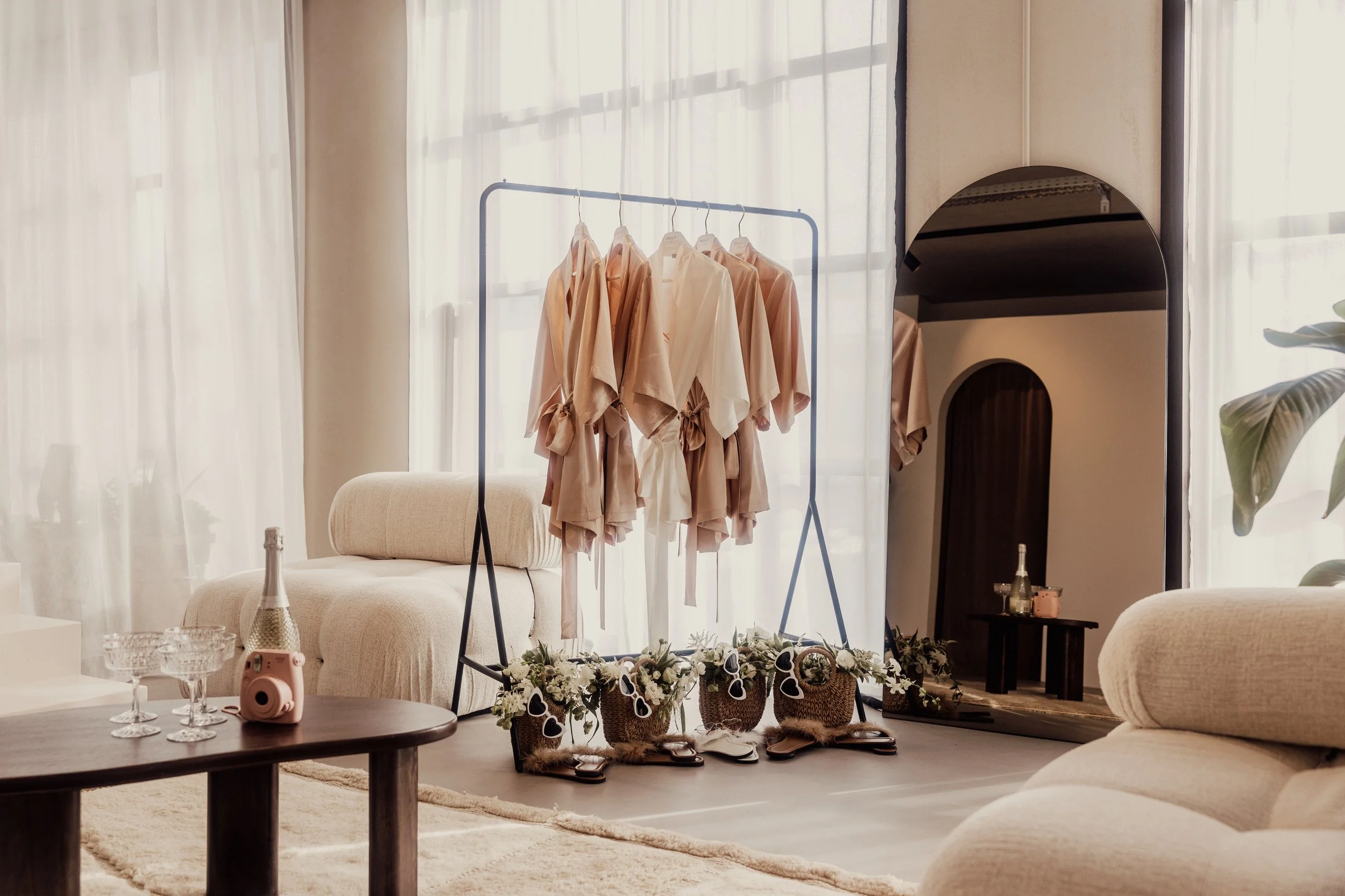 Interior scene with clothes rack holding beige robes, wicker bags with flowers and sunglasses, a round mirror, champagne bottle, glasses, and a camera on a wooden table, all in a softly lit room with beige furniture.