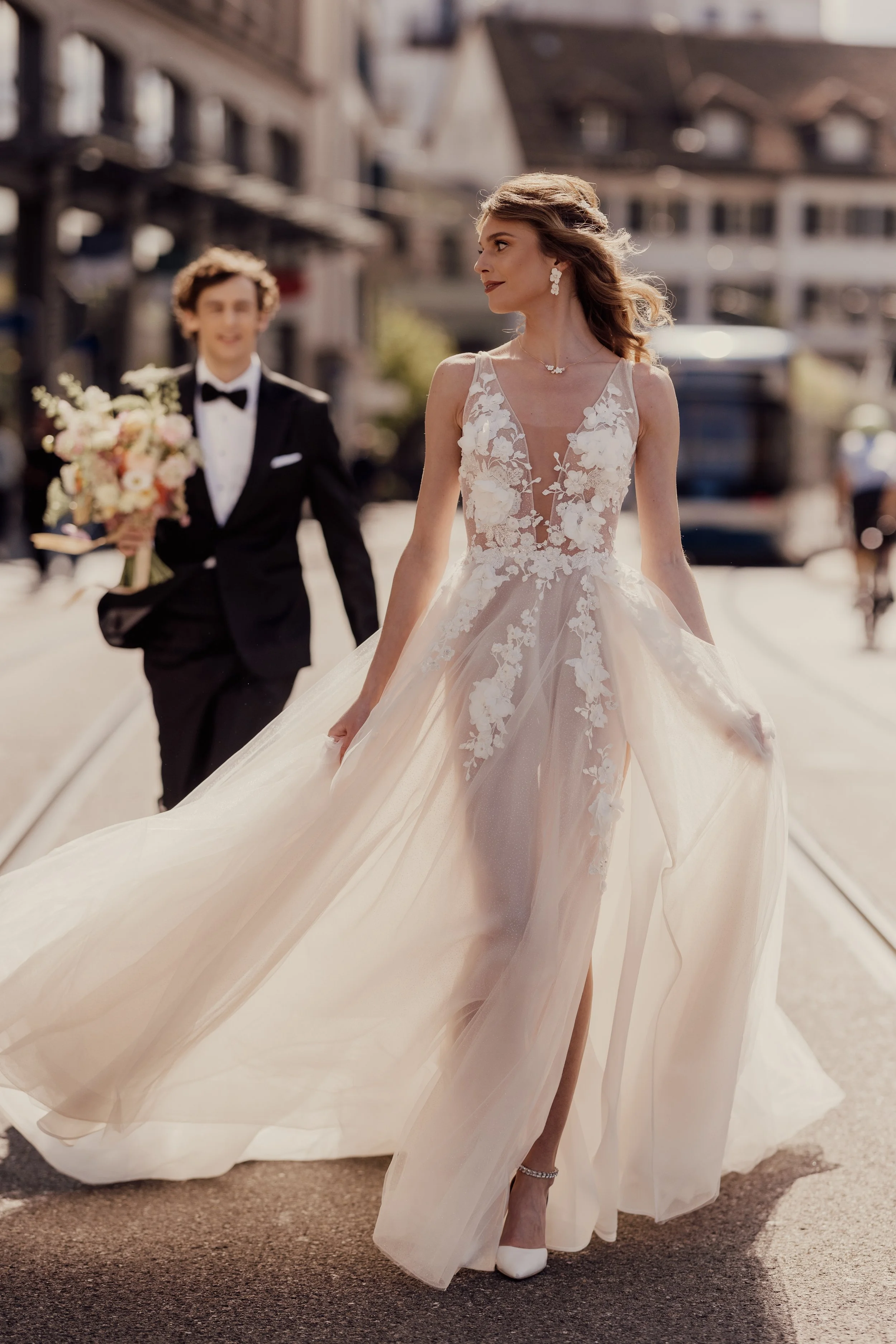 Bride in a flowing wedding dress walking down a city street, accompanied by a groom in a black tuxedo holding a bouquet.