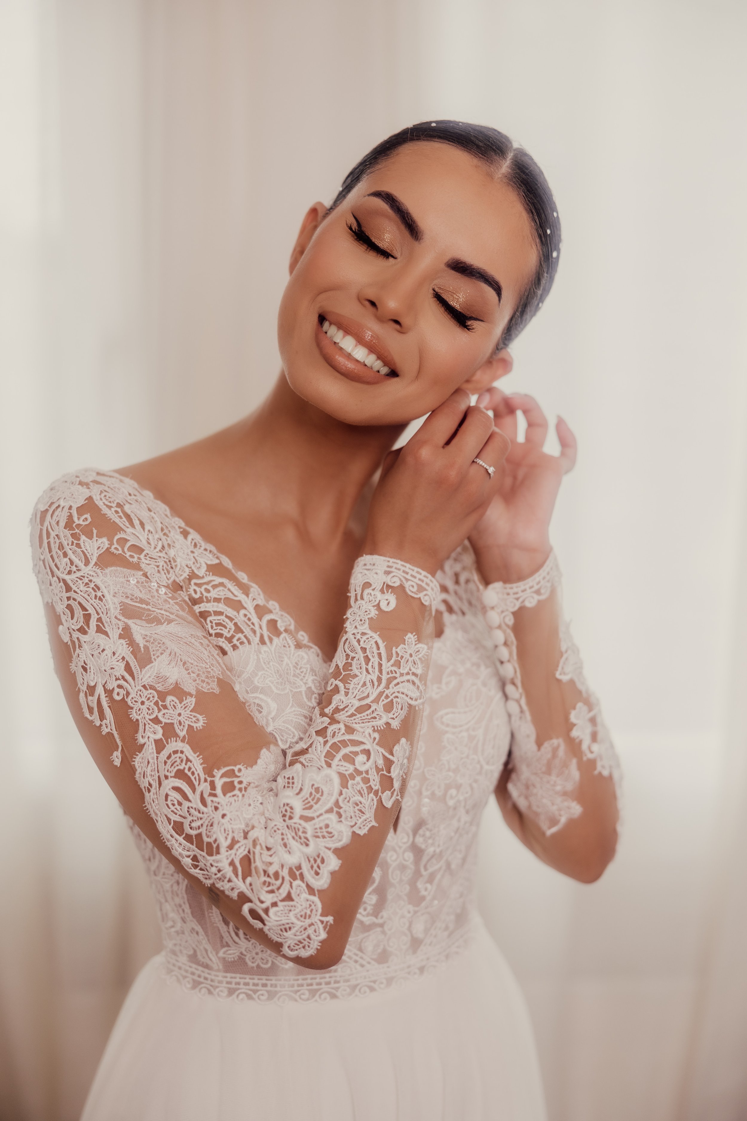 Bride smiling and wearing a lace wedding dress while adjusting an earring.