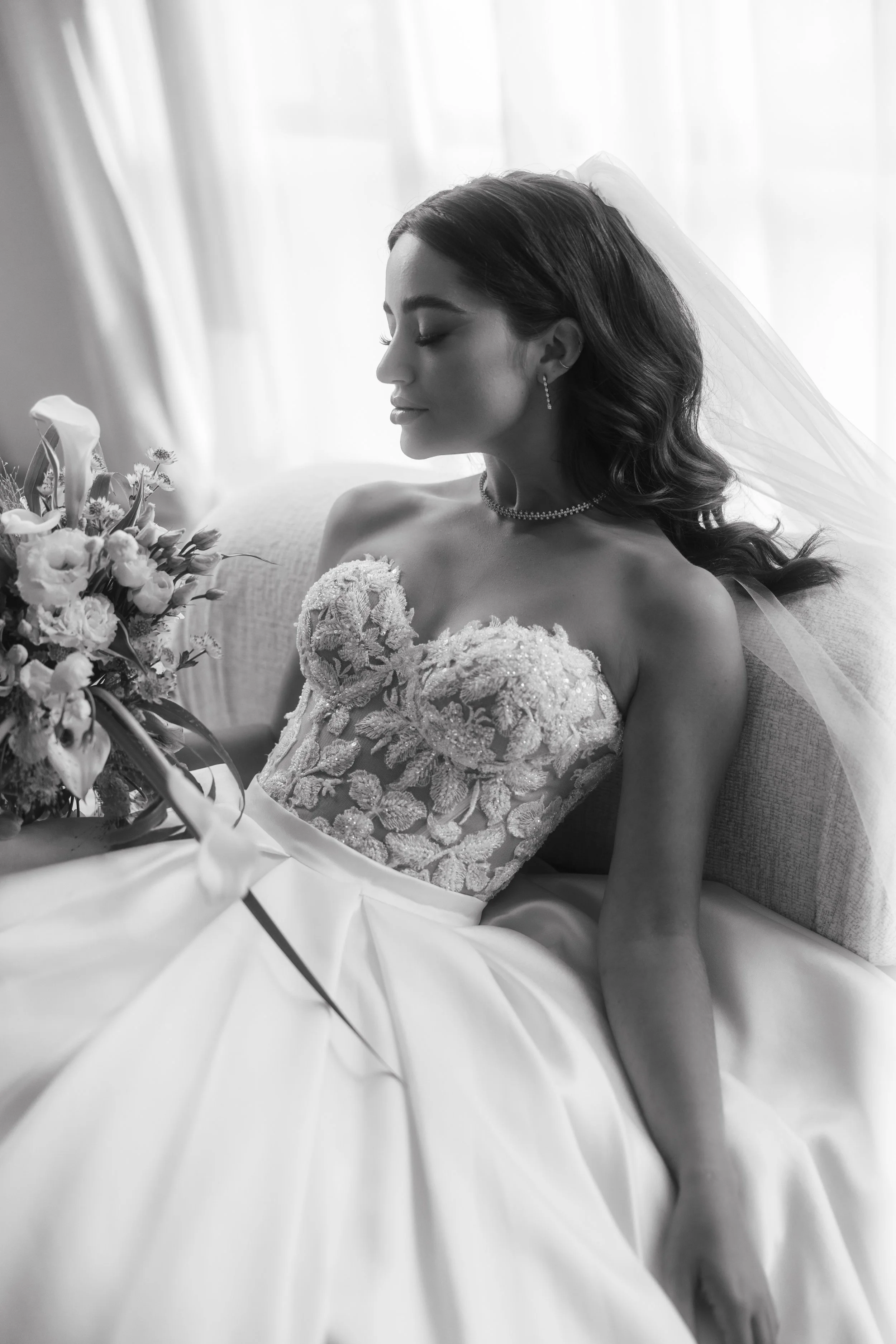 Black and white photo of a bride in a detailed lace bodice wedding dress, holding a bouquet. She is sitting with a veil over her hair, looking serene.