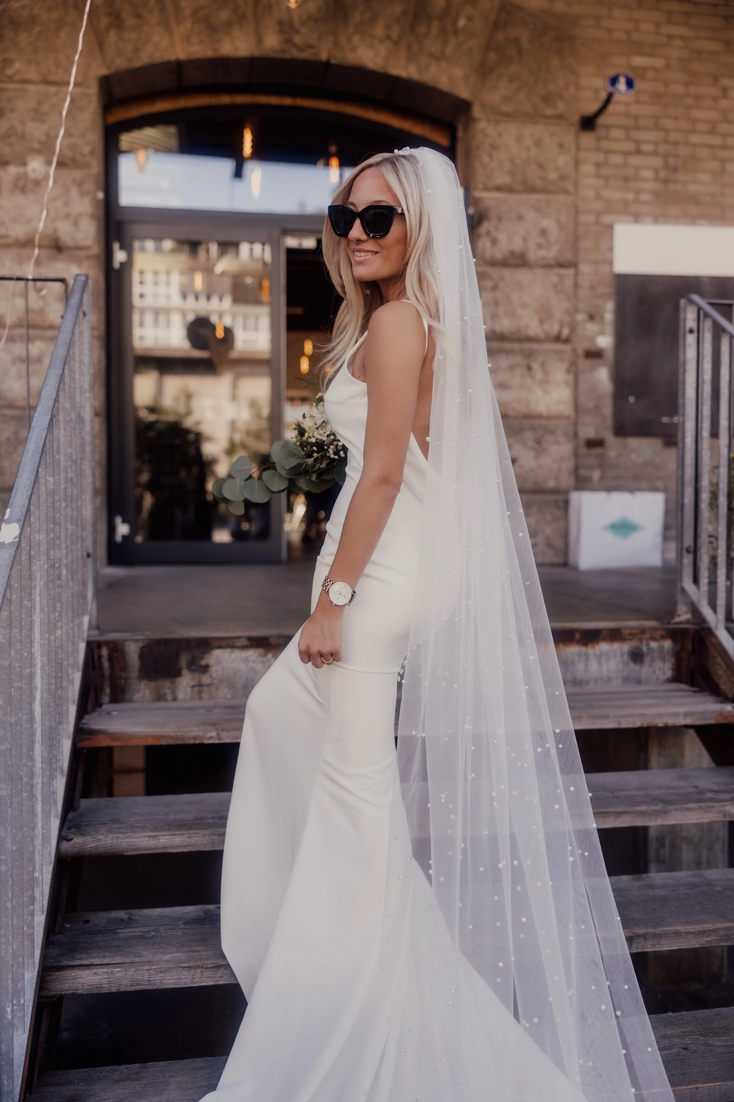Bride wearing a white wedding dress and veil, holding a bouquet, standing on stairs outside a brick building, wearing sunglasses.