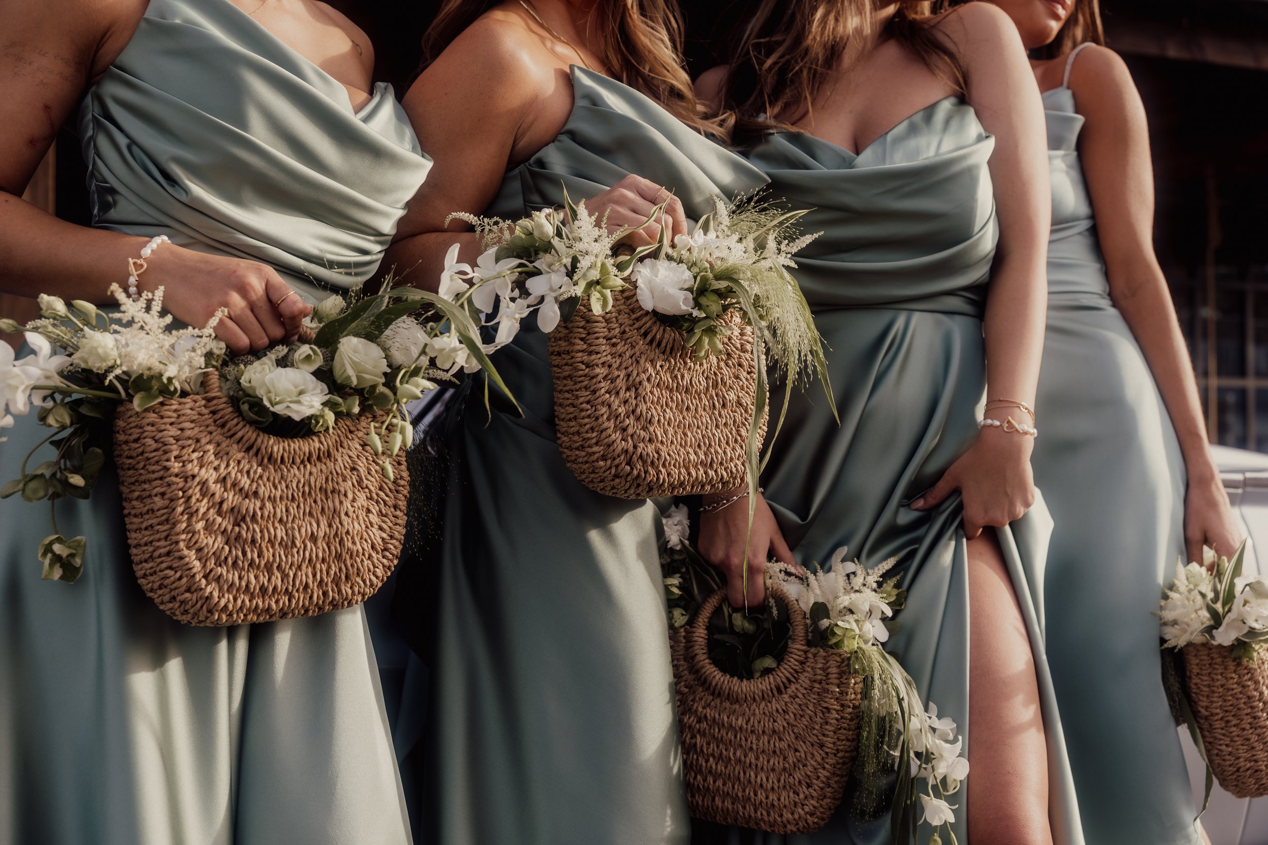 Bridesmaids in sage green dresses holding wicker baskets with white flowers.