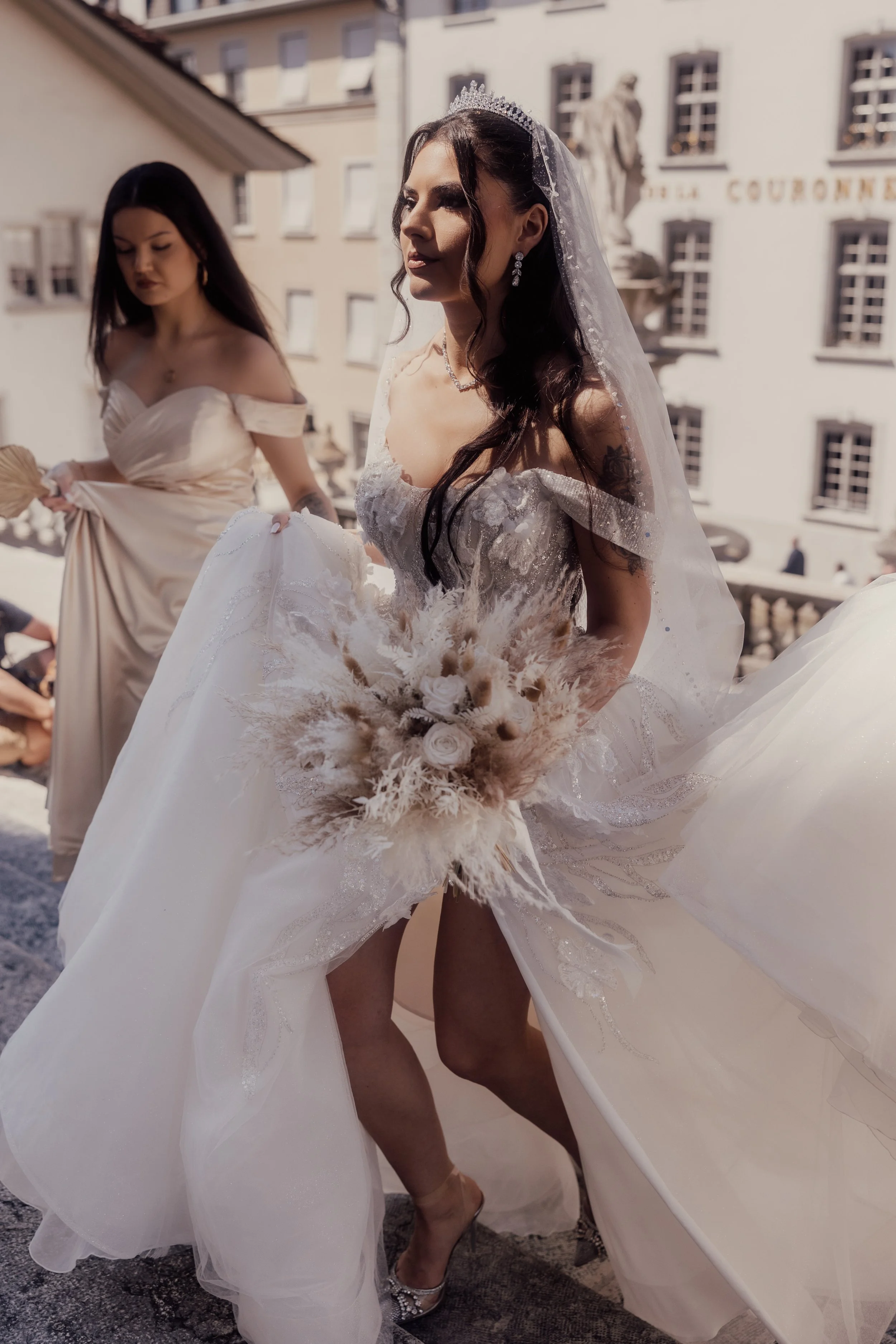 Bride in white wedding dress holding bouquet with a bridesmaid, outdoors, with buildings in the background.