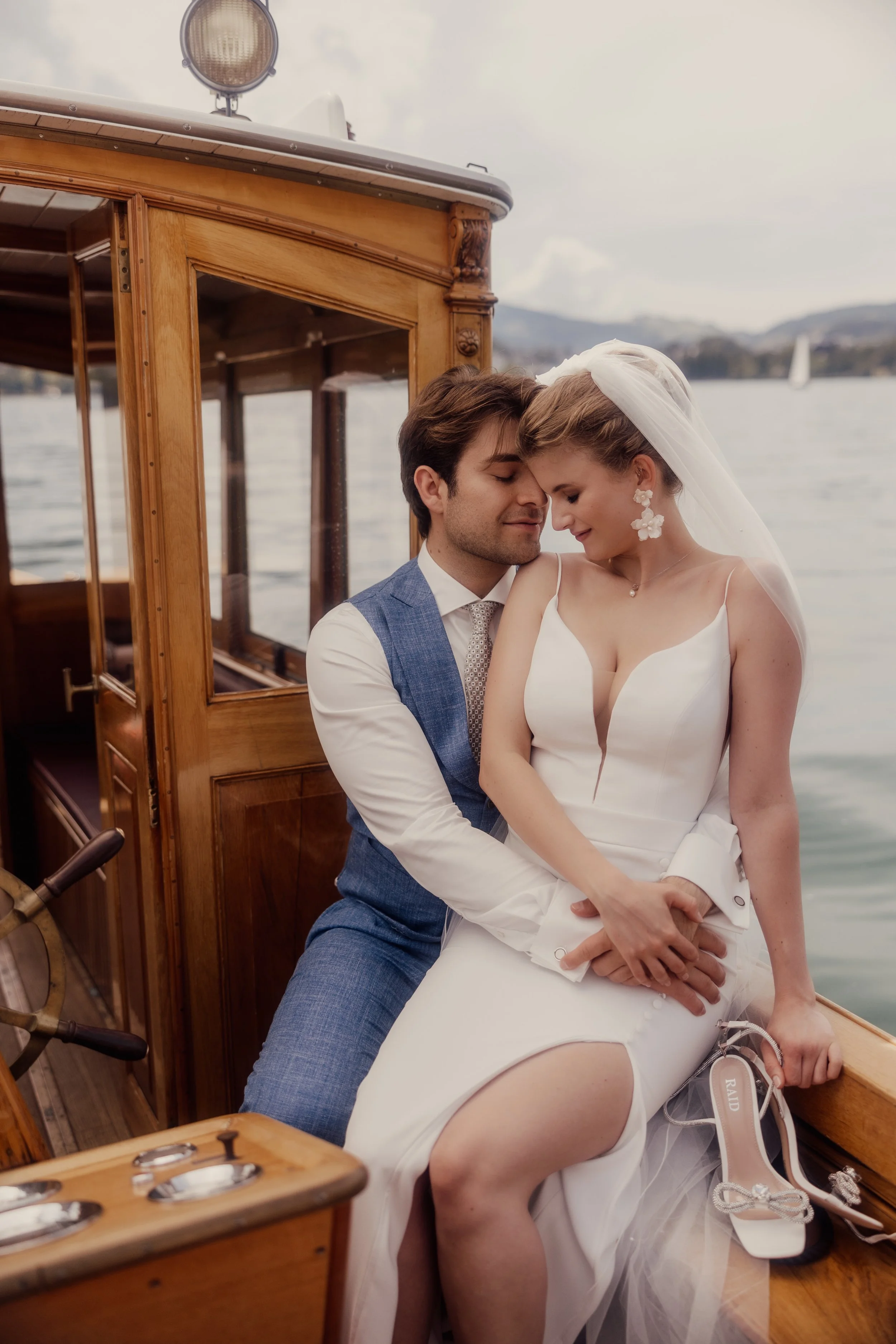 Bride and groom embracing on a wooden boat with water and mountains in the background, bride holding shoes, groom in blue suit.
