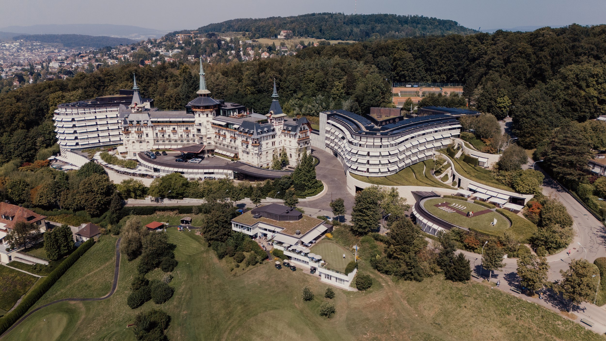Aerial view of a large luxury hotel complex with multiple connected buildings in a green, forested area, featuring landscaped gardens and nearby houses.