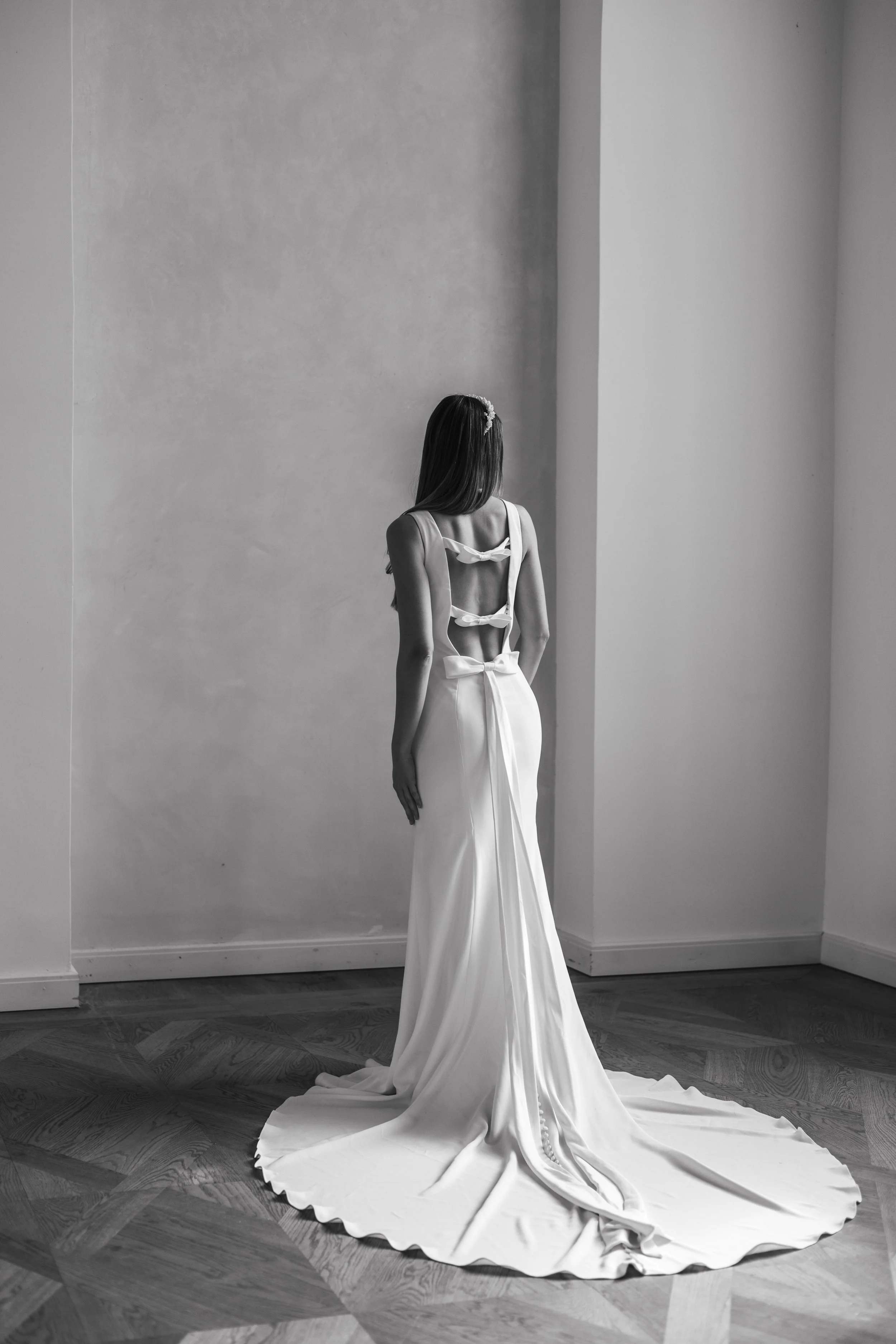 Black and white photo of a woman in a white bridal gown with an open back and bow details, standing indoors on a hardwood floor.