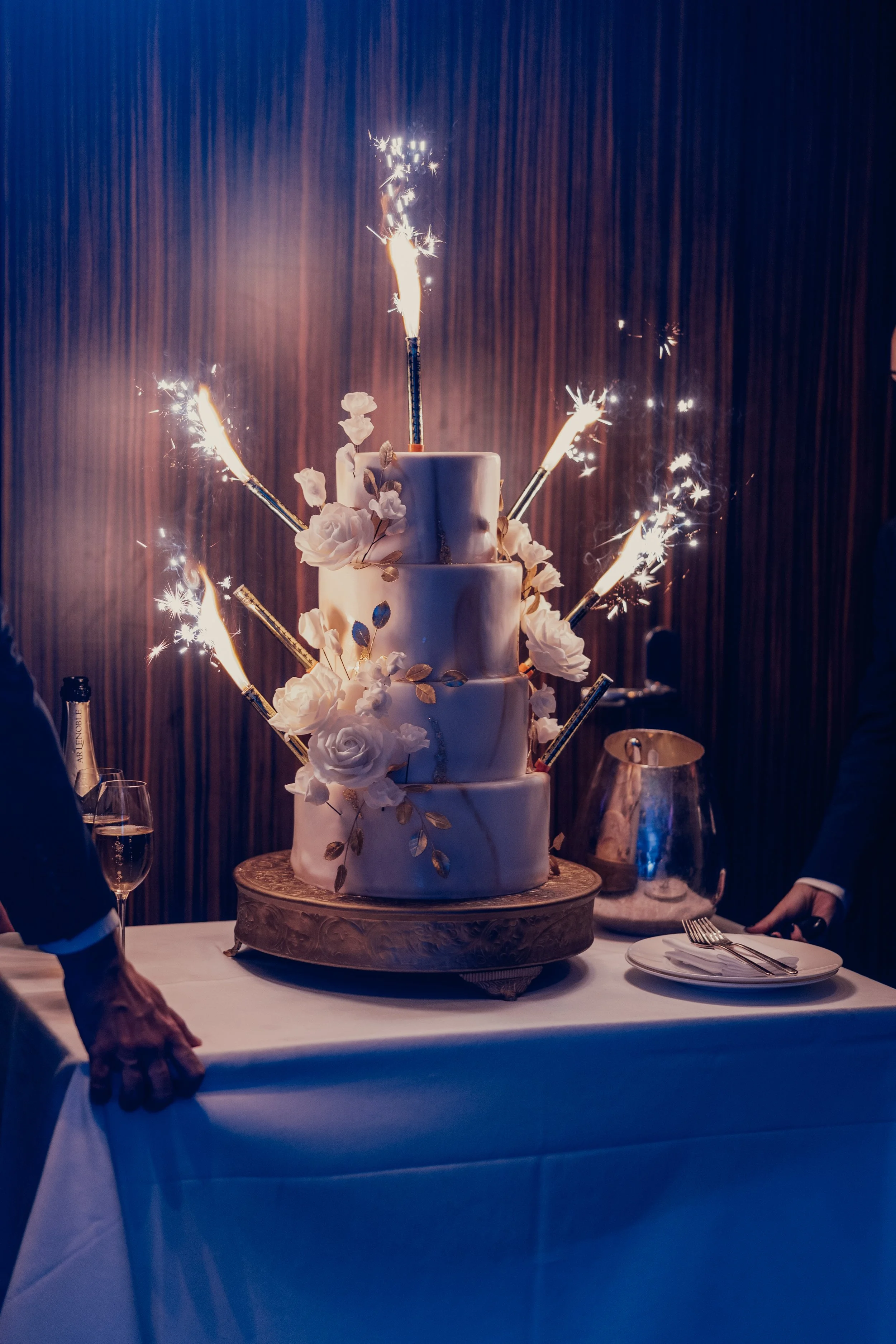 A three-tier white wedding cake adorned with white roses and gold leaves, with sparklers on top and around the cake, set on a decorative cake stand on a table.