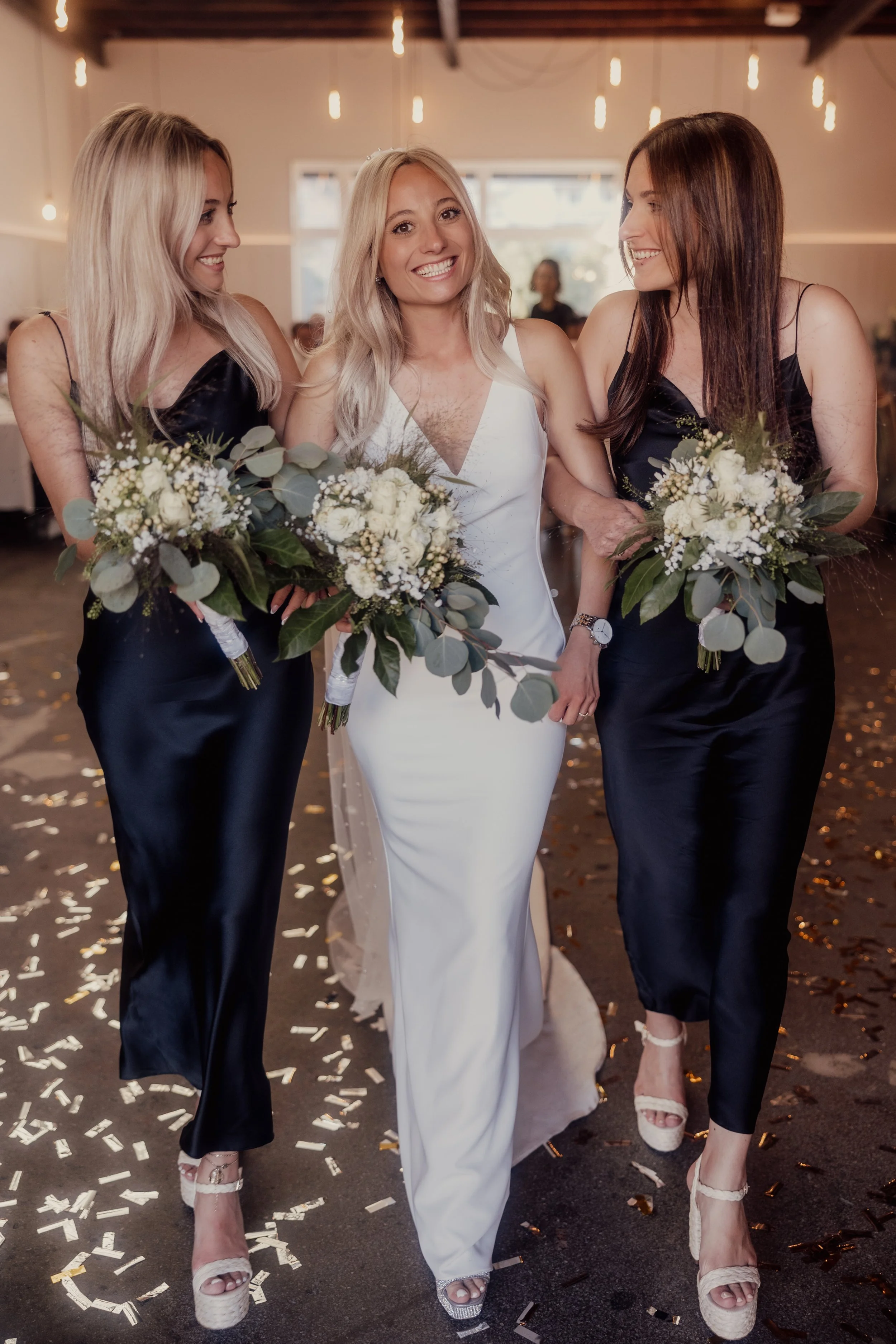 Bride in a white dress and two bridesmaids in black dresses holding bouquets, walking indoors, with confetti on the floor.