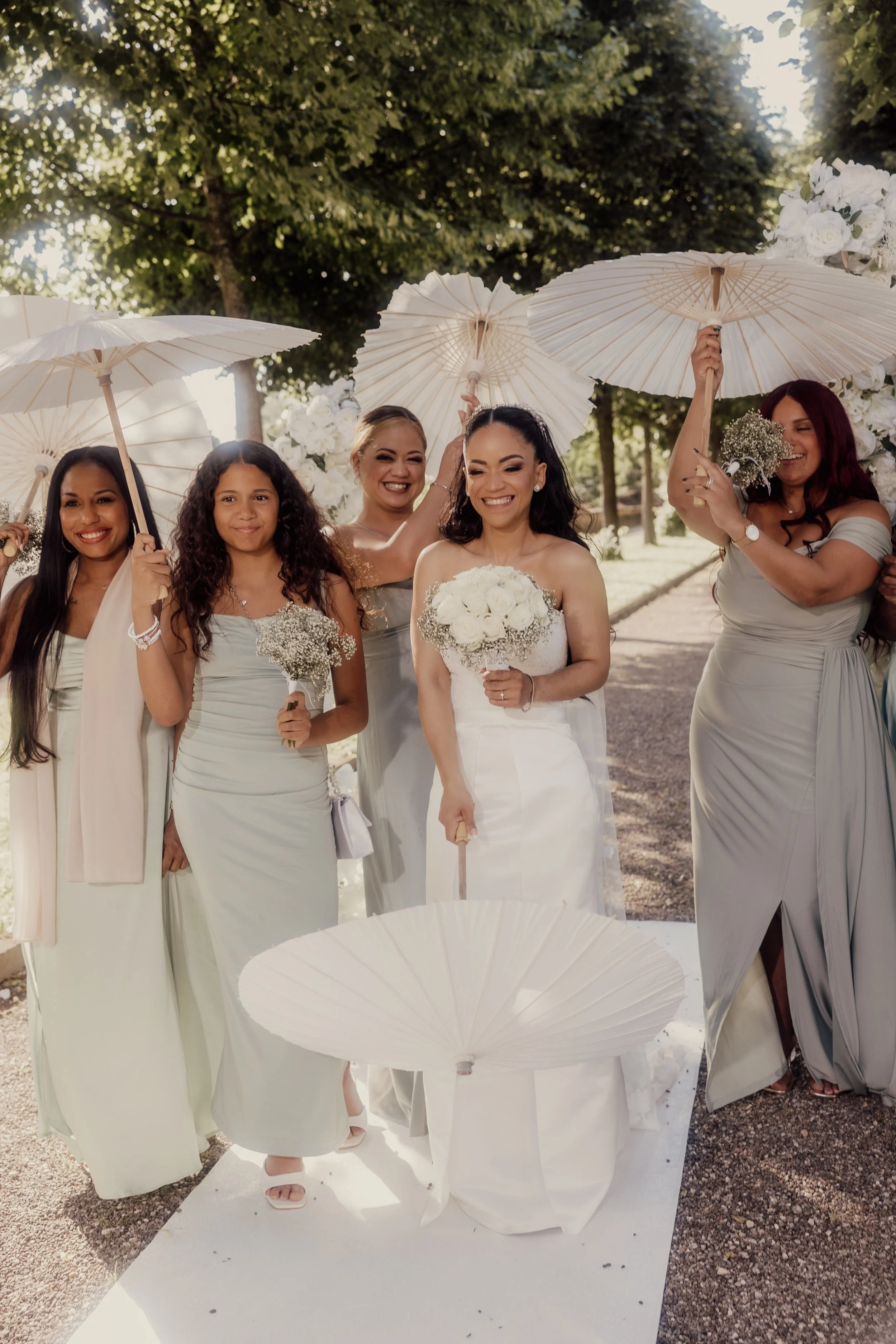 Bridal party with women in matching dresses holding parasols and flowers, standing on a white carpet outdoors.