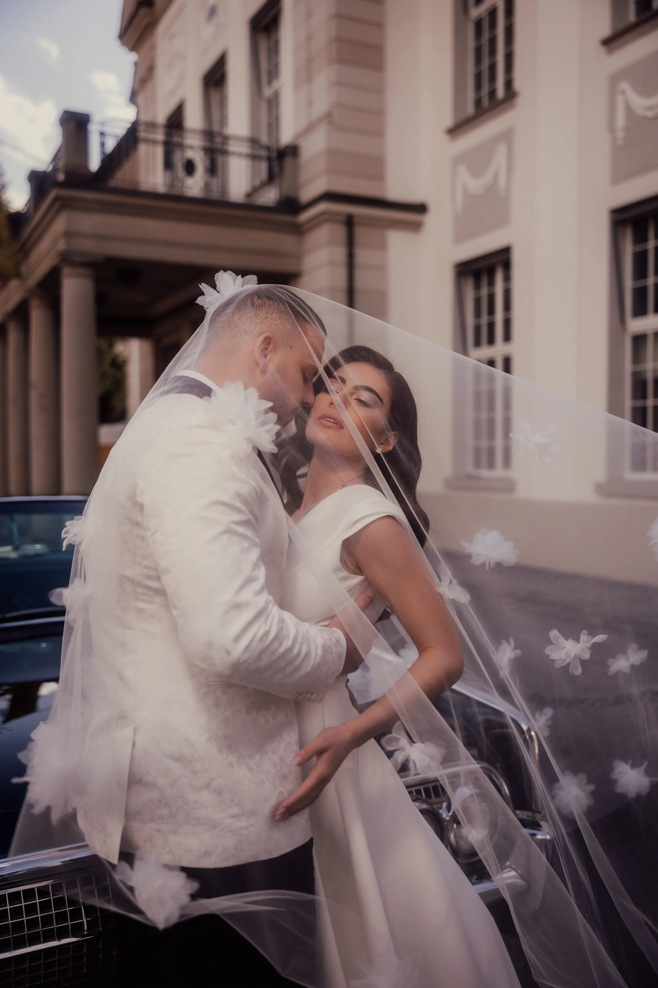 Bride and groom embracing under a veil in front of a classic car and elegant building.