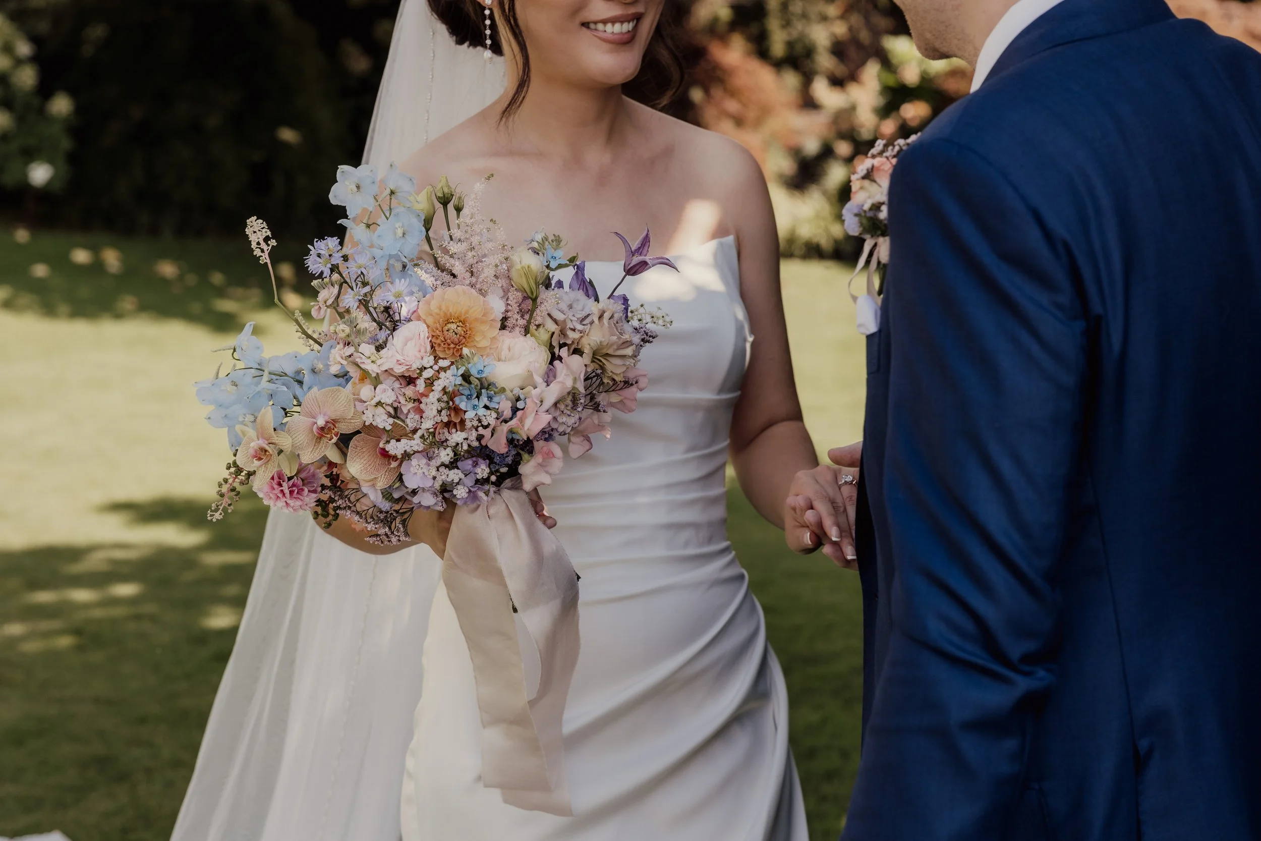 Bride holding a vibrant bouquet next to a groom in a blue suit, both standing outdoors during a wedding ceremony.