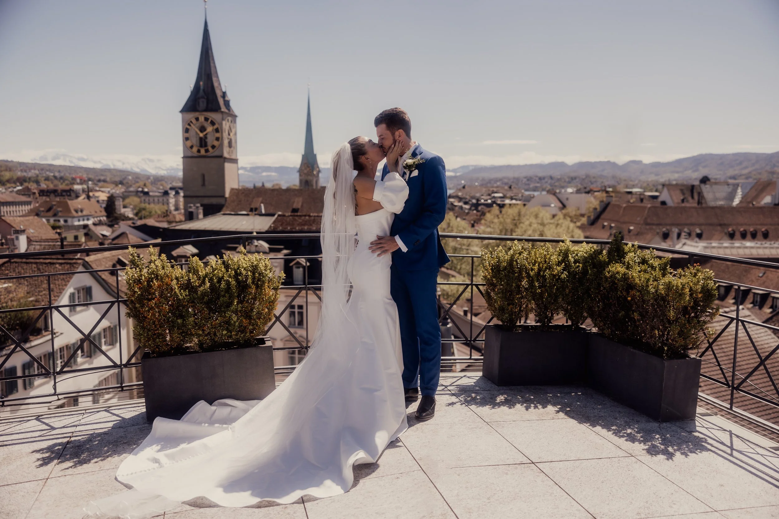 Bride and groom kissing on a rooftop with cityscape view, featuring church steeples and skyline in the background.