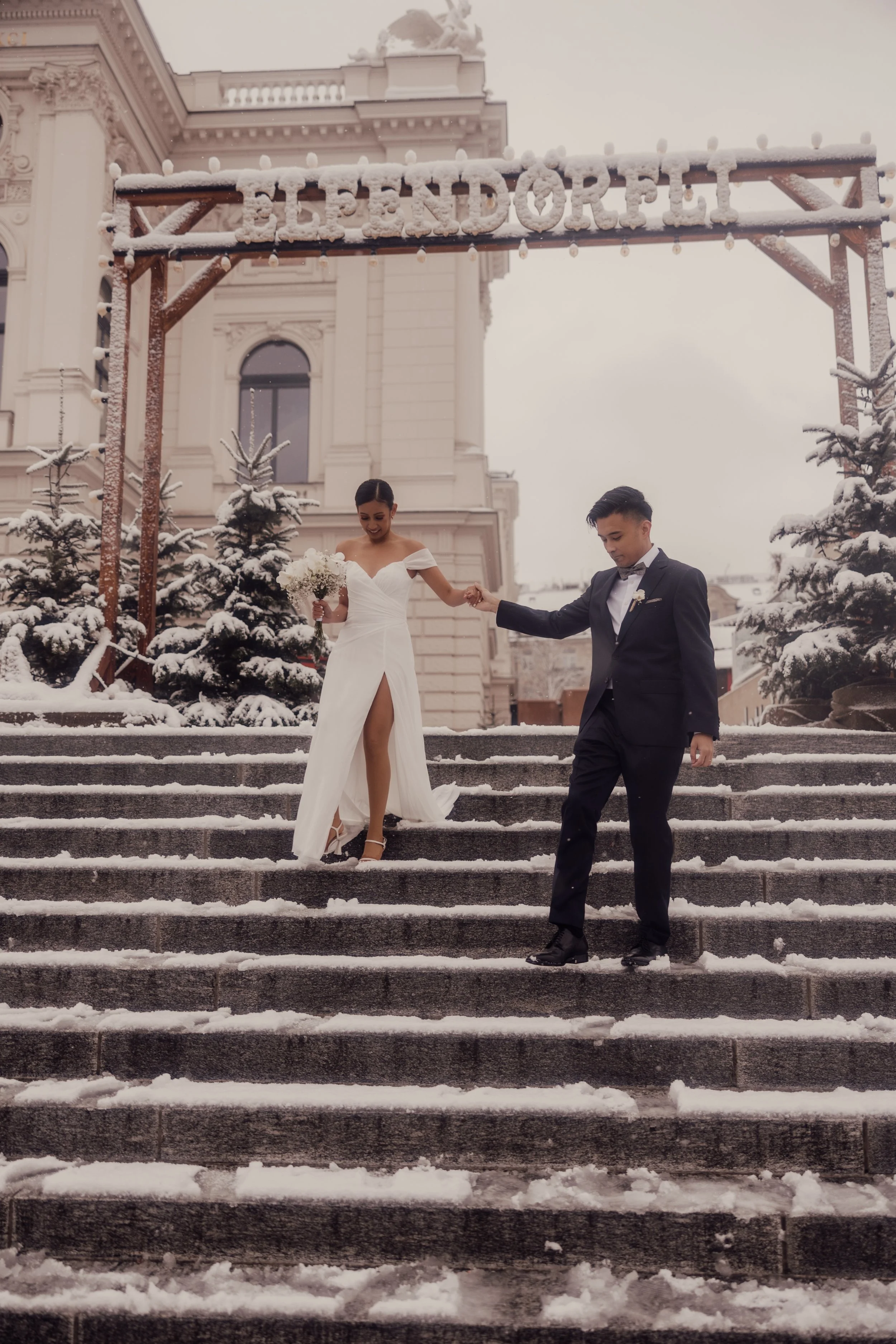 Bride and groom walking down snowy steps, bride holding a bouquet, groom wearing a suit, building in background.