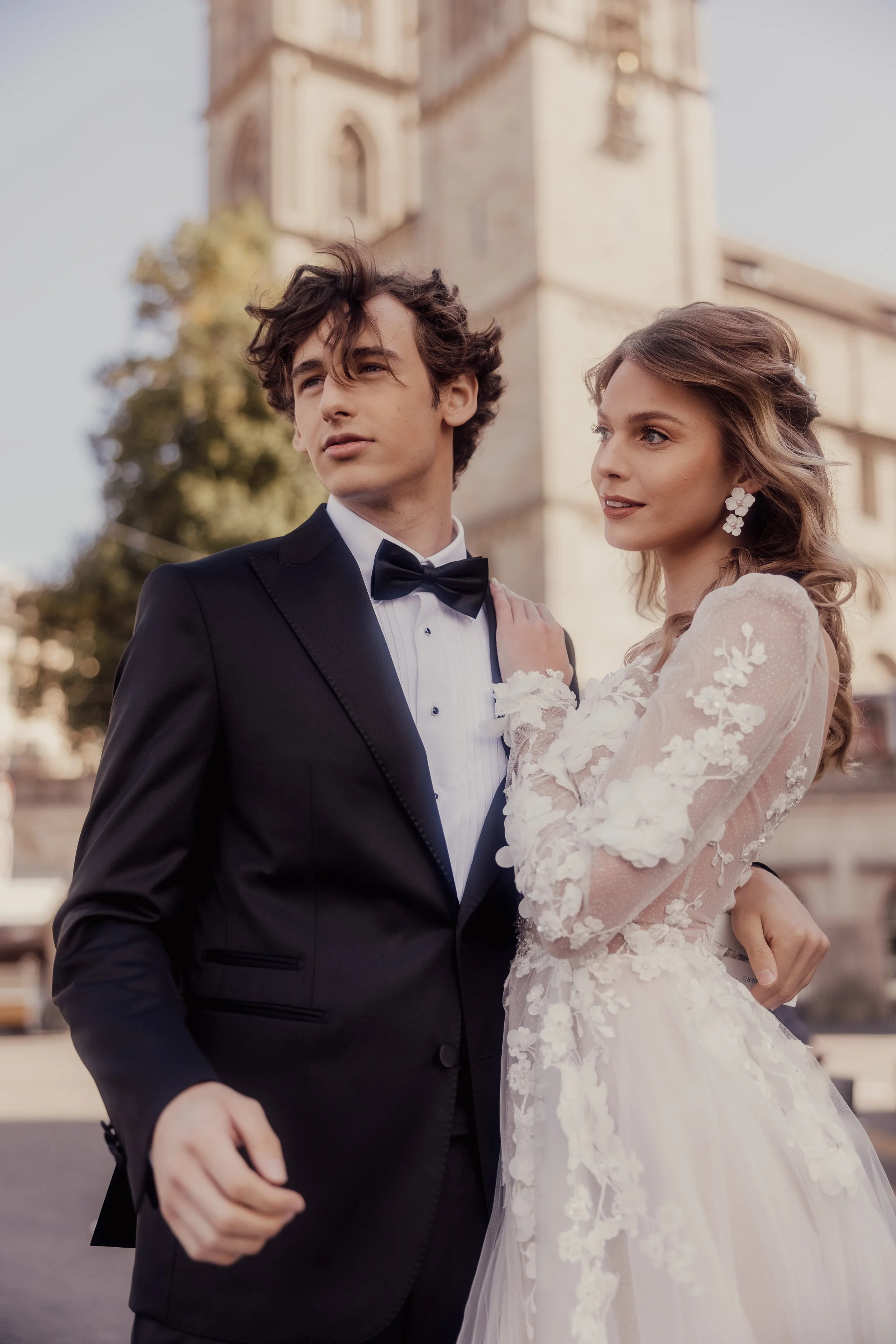 Bride and groom in elegant attire standing outdoors near a historic building.