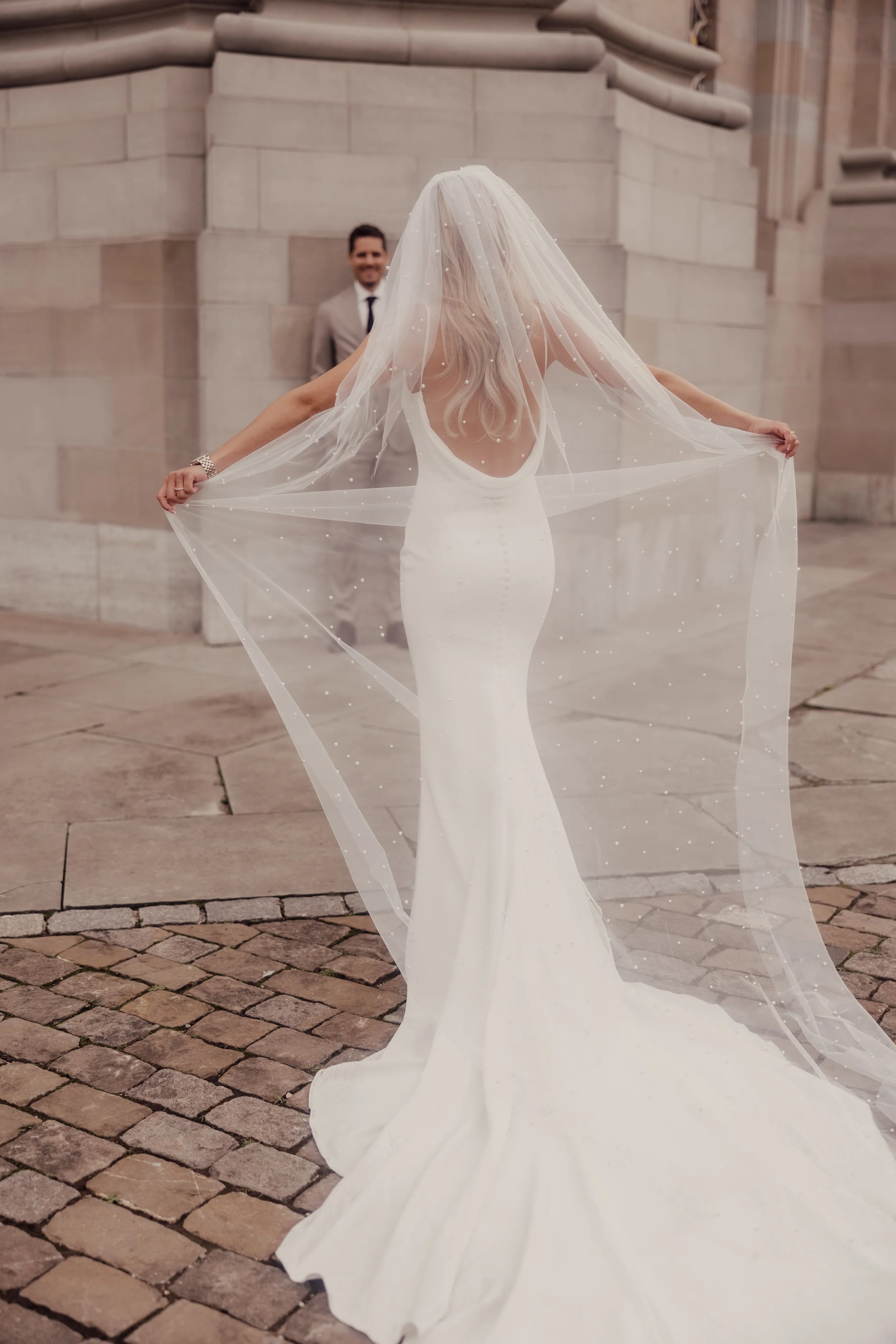 Bride in a long white wedding dress with a veil poses in front of a groom on a cobblestone street.